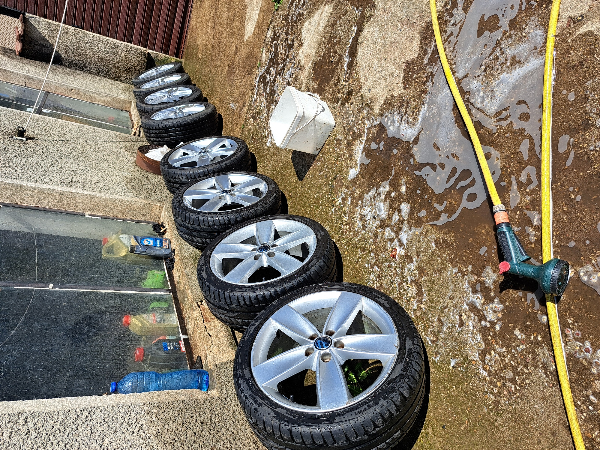 The image depicts a row of car wheels and tires lined up on a concrete surface next to a wall. The tires appear to be used, and some have a silver rim. There is also a yellow water hose with a spray nozzle lying on the ground, with soapy water on the surrounding concrete. A white bucket and various bottles are present nearby. The location is identified as Chepintsi, Bulgaria.