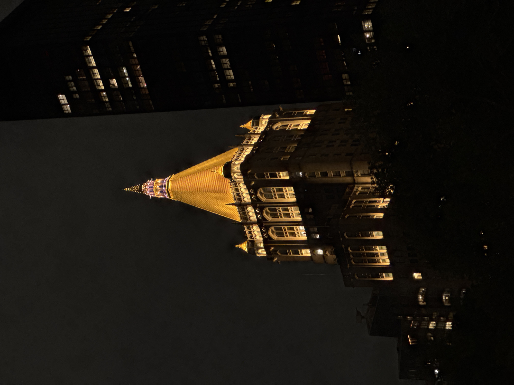 The image is a nighttime view of the New York Life Building, located in New York City, USA. The building's distinctive gold-leaf pyramid-shaped tower is illuminated, as are many of its windows. The surrounding sky is dark, and the presence of adjacent buildings and possible trees suggests an urban setting. The details of the windows indicate a multi-story building.
