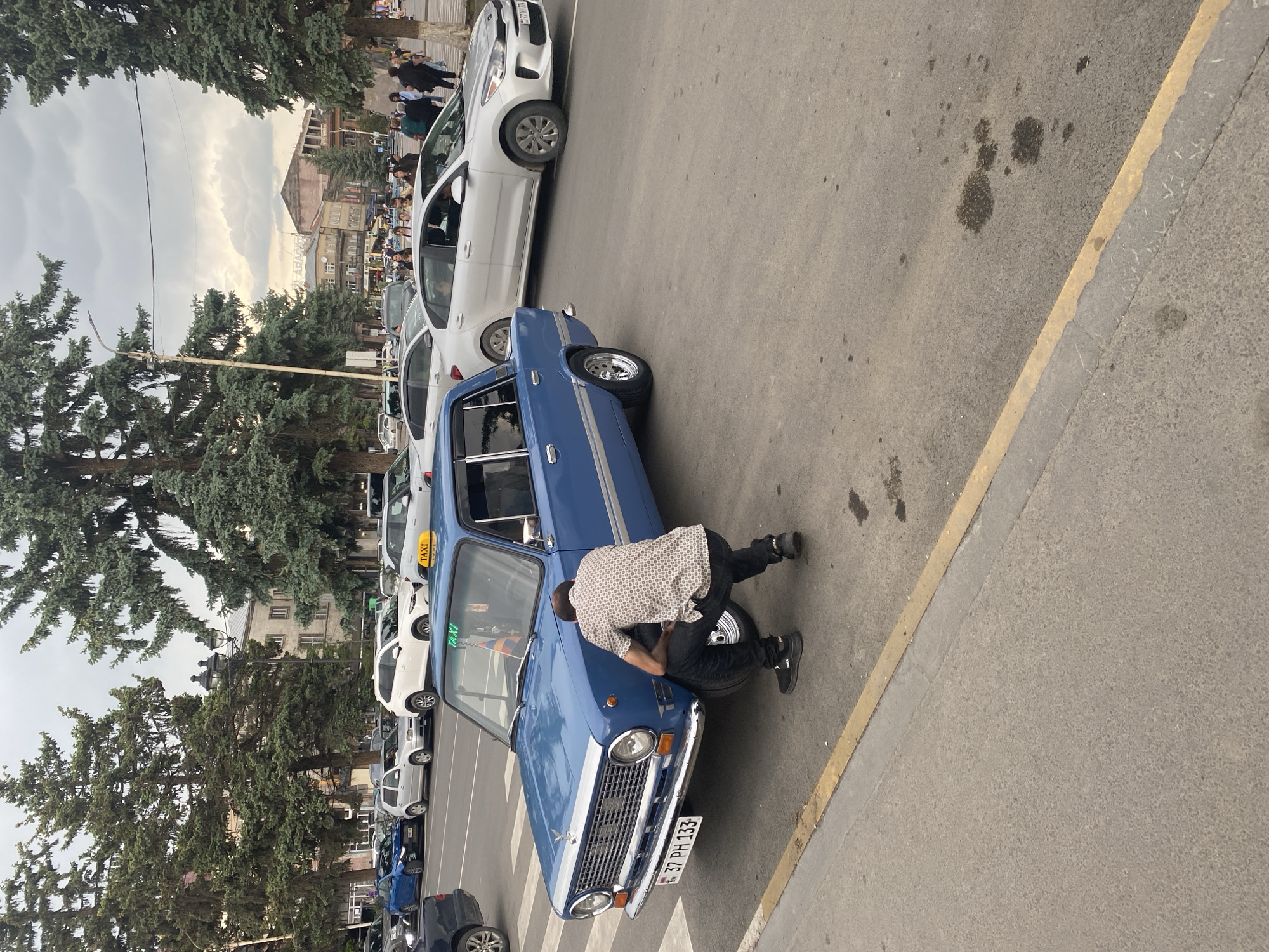 A man in a patterned shirt and dark pants is crouched down, appearing to adjust the front wheel of a blue vintage taxi on a paved street in Gyumri, Armenia. The taxi has a yellow "TAXI" sign on its roof and an Armenian flag decal on the windshield. Other vehicles, including a white sedan with a "TAXI" sign, are visible in the background, along with trees and buildings, suggesting an urban setting. The sky is overcast.
