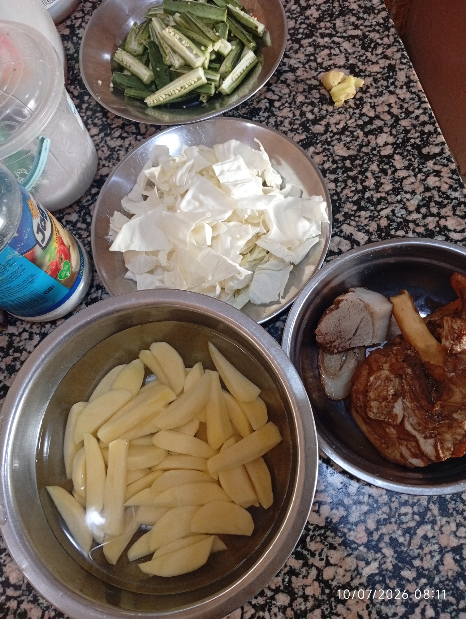 In Bana Stream, Nigeria, a close-up, top-down view captures ingredients for a meal laid out on a granite countertop. In a stainless steel bowl on the lower left, chopped potatoes are submerged in water. To the right, another stainless steel bowl contains cooked meat, appearing to be beef or lamb, with visible fat and bone. Above and slightly to the left, a silver plate holds chopped cabbage. Further up, a similar plate displays sliced okra. A small piece of ginger rests on the countertop between the okra and cabbage. To the left, a partially visible plastic container filled with a white granular substance, possibly flour or sugar, and a bottle with a blue label are also present. A timestamp at the bottom right corner reads "10/07/2026 08:11".