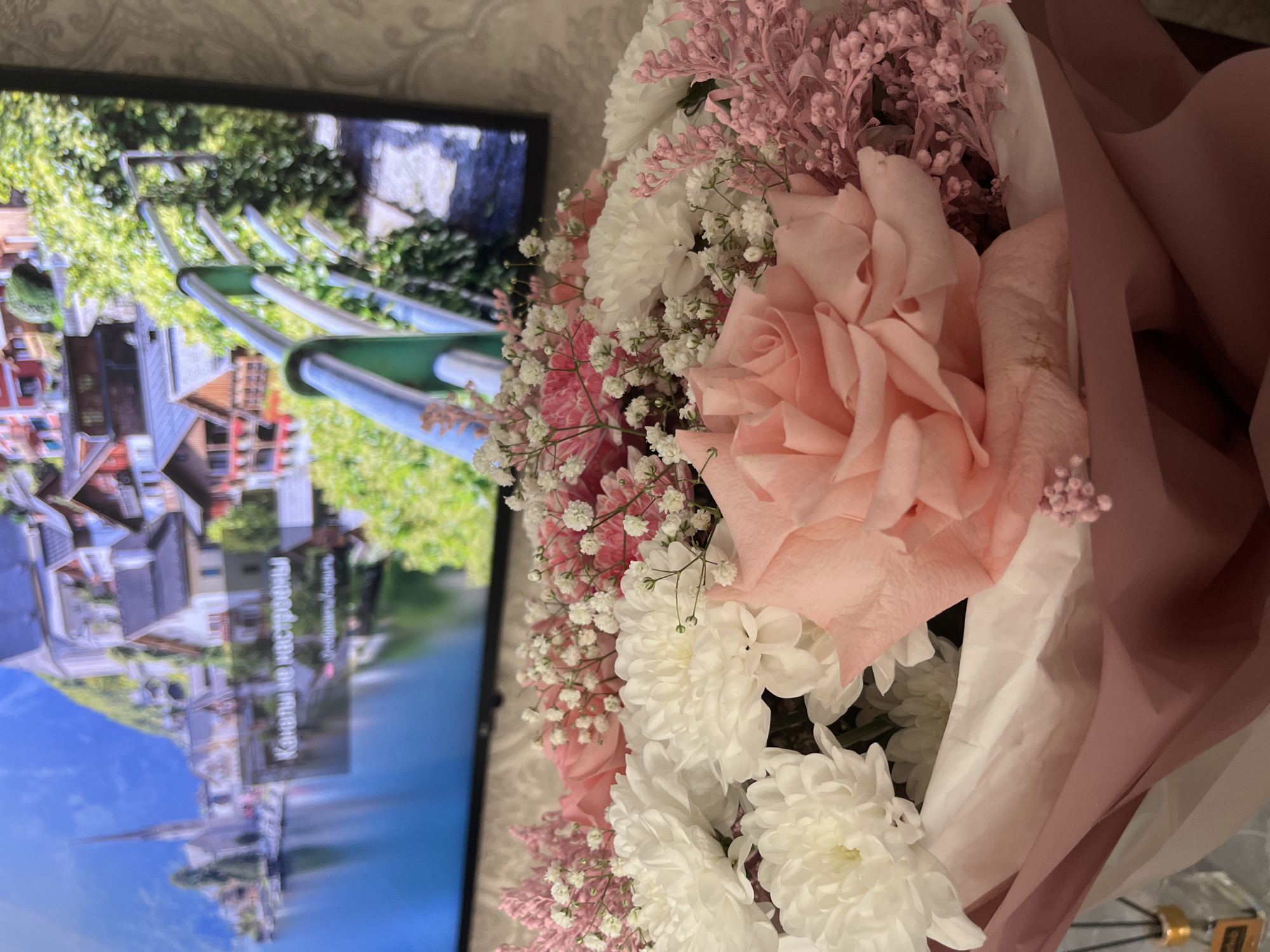 The image shows a bouquet of flowers in the foreground, with a television screen in the background displaying a scenic view of Hallstatt, Austria. The flowers include a large pink rose, white chrysanthemums, baby's breath, and pink filler flowers. The bouquet is wrapped in light pink and white paper. The TV screen shows a village with traditional Austrian architecture along a lake, with a green pedestrian bridge spanning the water. Text in Russian is visible on the screen. The location of the image is not discernible from the image itself.