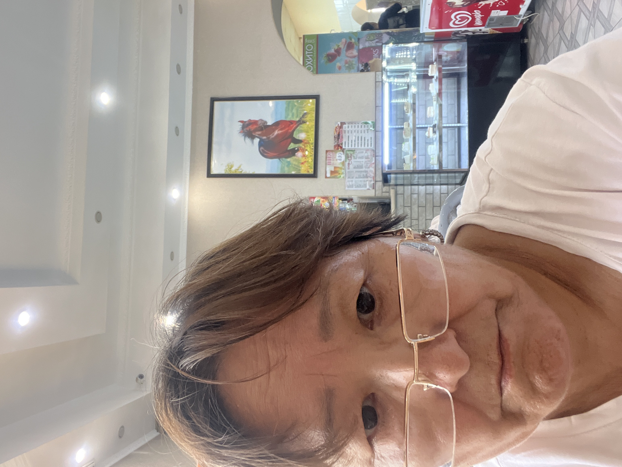 A close-up, front-facing selfie shows a woman in her 50s or 60s with brown hair and glasses, looking directly at the camera with a neutral expression. She is wearing a white t-shirt. In the background, there's a display case filled with cakes, a framed picture of a brown horse, and what appears to be an ice cream freezer. The scene suggests an indoor environment, possibly a cafe or dessert shop. The overall setting is consistent with a location in Petropavl, Kazakhstan, given the text visible on some of the items in the background.