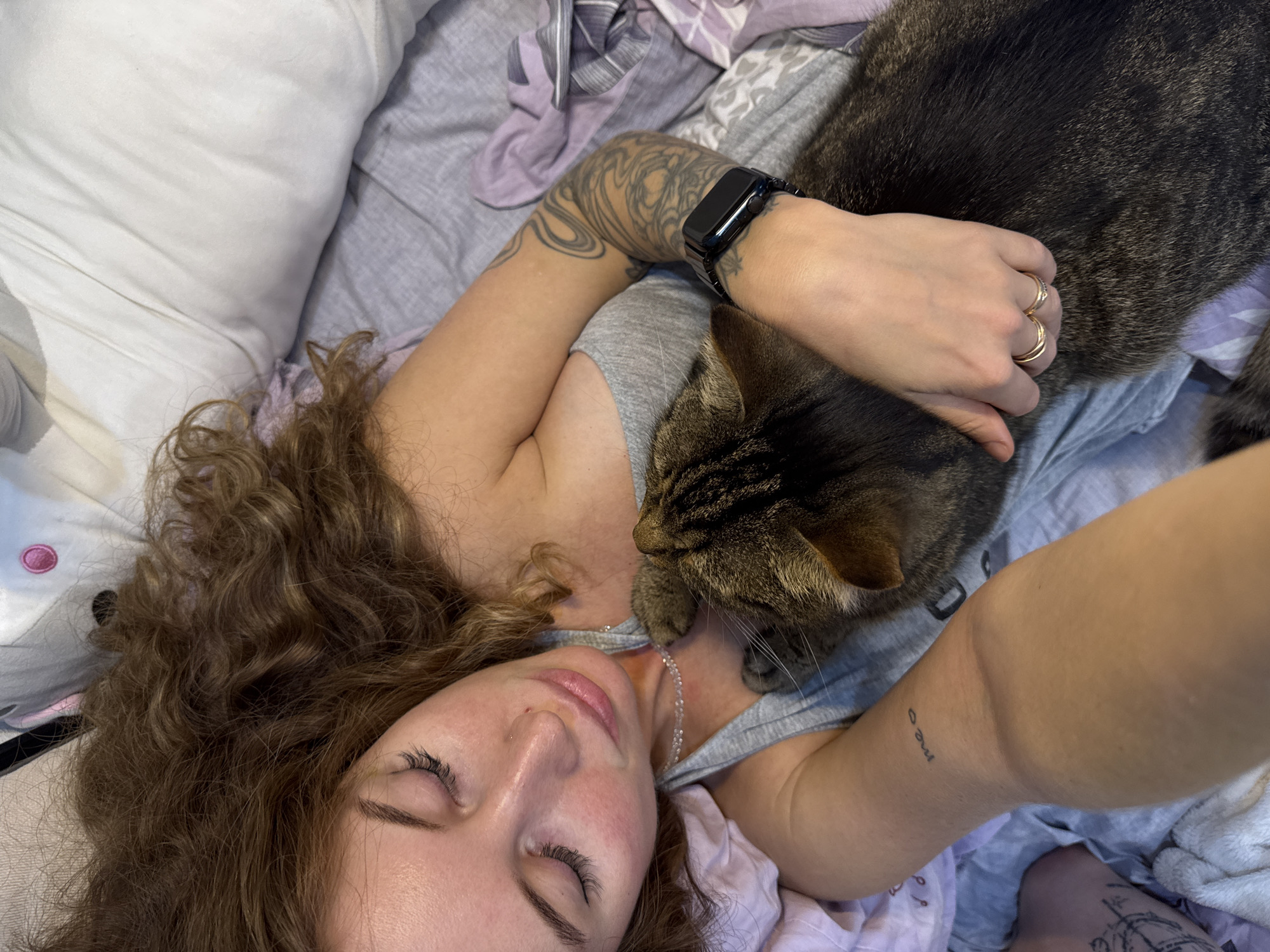 The image depicts an indoor scene in the city of Chaikovsky, Russia. A light-skinned woman with long, curly brown hair is shown lying on her back in a bed, with her head turned slightly to her left. Her eyes are closed, indicating sleep or deep relaxation. She is wearing a grey tank top and a delicate, clear beaded necklace. A small stud piercing is visible on her left nostril. Tattoos are prominent on her right arm, forming a full sleeve of intricate black linework designs. A smaller tattoo of the word "meo" is present on her left forearm. A black smartwatch with a dark metal band is worn on her right wrist, and two rings (one plain gold, one with small stones) are on her left ring finger.
A large, dark brown tabby cat is nestled against the woman's chest and shoulder area, its head turned towards her face. The woman's left arm is gently draped over the cat's back. The cat appears relaxed, possibly sleeping. The bedding consists of light grey and purple patterned sheets, with a white pillow visible under the woman's head. Additional crumpled purple fabric is present on the bed.