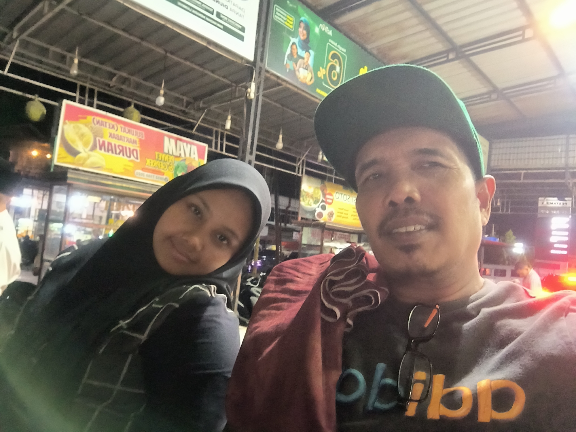 A nighttime selfie shows two individuals in front of a food stall in Meukokuthang, Indonesia. On the left is a woman wearing a black hijab and a patterned dark blue shirt. To her right is a man wearing a green baseball cap and a dark grey t-shirt with yellow lettering. He has sunglasses hanging on the front of his shirt and a maroon cloth draped over his shoulder. Various food stalls are visible in the background, with signs advertising durian and other foods. Lights are strung overhead. A building is visible across the street, with illuminated vertical signs. Other people are in the background, blurred due to the camera focus.