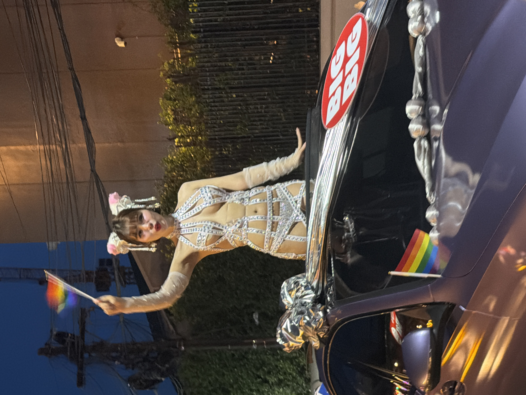 The scene depicts a person, presumably participating in a parade or celebration, standing next to a car decorated with rainbow flags and shiny ornaments. The person is holding a small rainbow flag and is wearing a white, sparkly outfit with matching gloves and headwear. The car has a "BIG" sign on it. The location appears to be an urban setting, likely Chiang Mai City Municipality, Thailand, given the presence of power lines, foliage, and street lights. The sky is dark, suggesting it is evening or night.