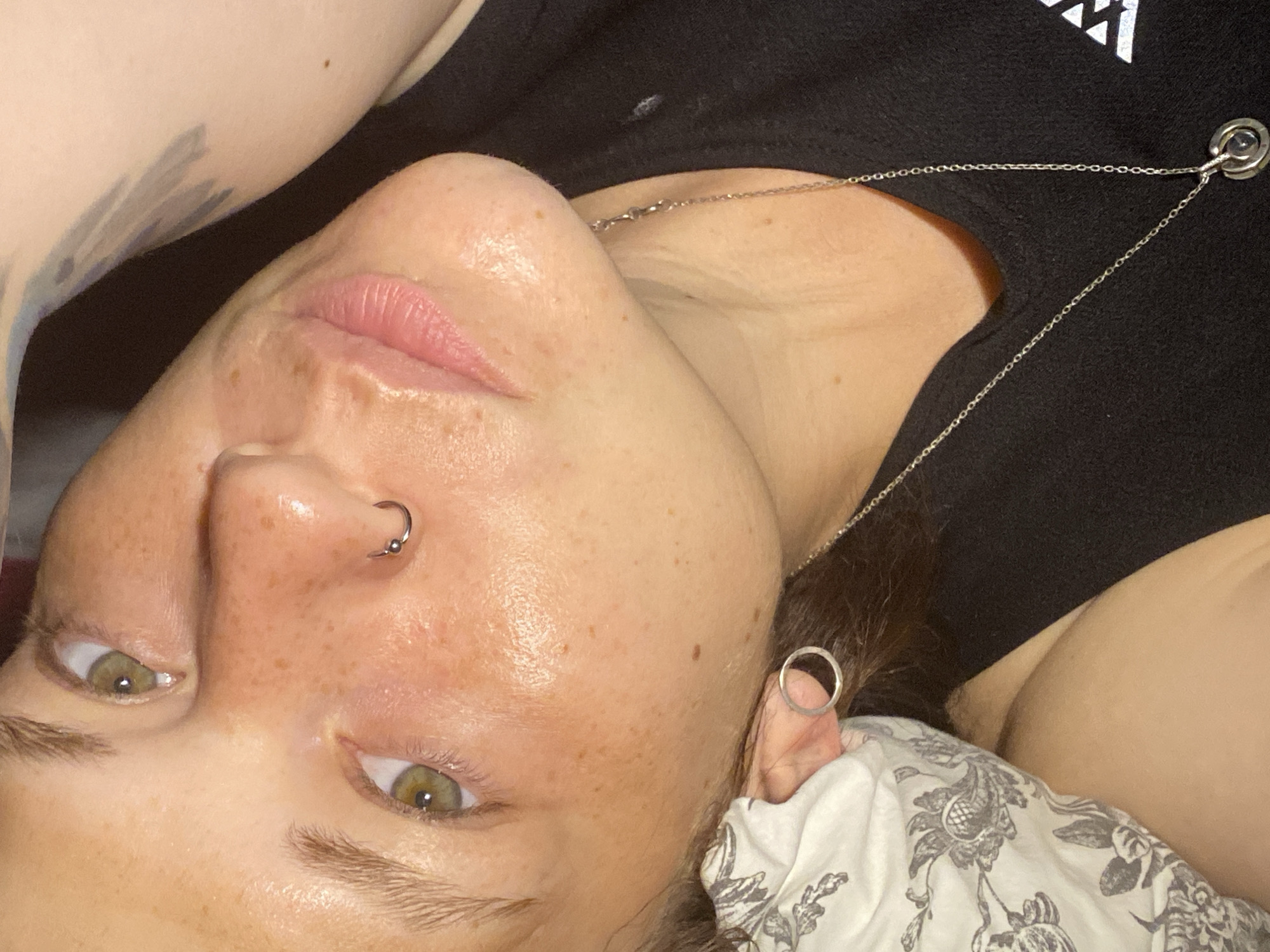 A close-up, head-and-shoulders shot shows a person lying on their side, facing the camera. The person has fair skin with freckles, green eyes, and light brown eyebrows. They are wearing a silver hoop earring in their left ear and a silver necklace with a circular pendant. The person is also wearing a black t-shirt. The background includes a floral patterned pillow on the left and a dark background on the right, which appears to be a blanket or clothing. A tattoo is partially visible on the upper right arm. The image quality is slightly grainy, and the lighting is soft, creating a warm glow on the skin. The exact location in Chaikovsky, Russia, cannot be determined from the visual information provided.