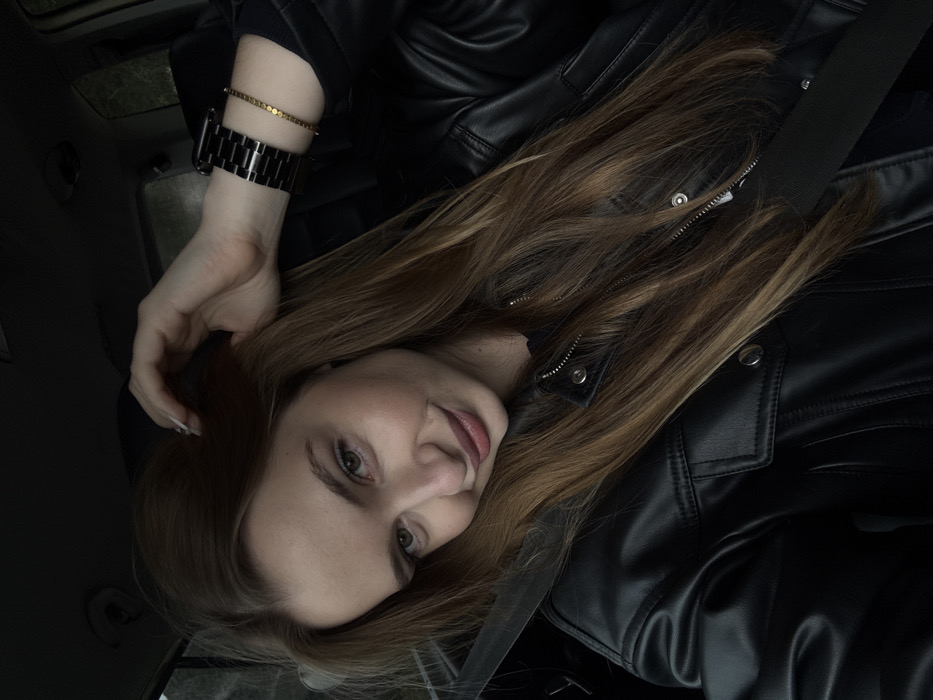 A female individual is depicted seated in the passenger side of a vehicle in Stupovi, Montenegro. She has long, light brown hair parted on the left, and light-colored eyes. Her head is tilted slightly, and she displays a subtle smile, looking directly at the viewer. Her left hand, with light-tipped fingernails, is raised and touches her hair above her right ear. On her left wrist, she wears a black metal link watch and a thin gold-colored bracelet composed of small, flat circular elements. She is dressed in a black leather-style jacket over a dark inner garment. A black seatbelt is visible, extending diagonally from her right shoulder across her chest and torso. The vehicle's interior serves as a dark, indistinct background, with faint indications of an exterior view through the windows.