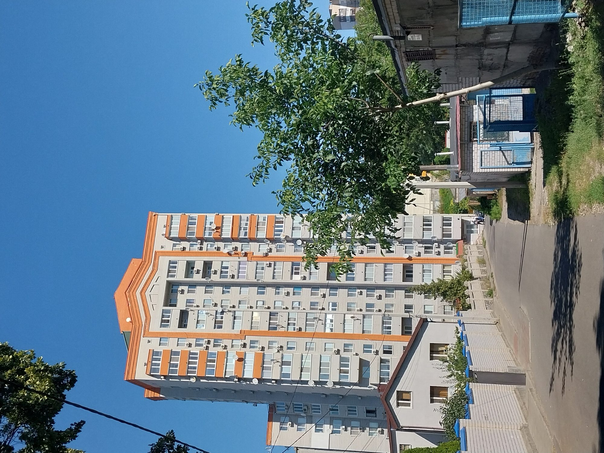 In Doslidne, Ukraine, a wide, paved road curves gently uphill, bordered by a white brick wall topped with blue decorative elements on the left. Beyond the wall is a low-rise building with a gabled roof. A tall, multi-story apartment building with orange trim and numerous windows dominates the background. To the right of the road are a series of low, utilitarian structures, including a brick building with blue metal gates and a corrugated metal structure with an open doorway. A leafy green tree stands prominently in the foreground on the right, its branches reaching towards the sky. The sky is a clear, bright blue. Shadows from the tree are cast on the road. Two individuals are walking in the distance on the left side of the road.