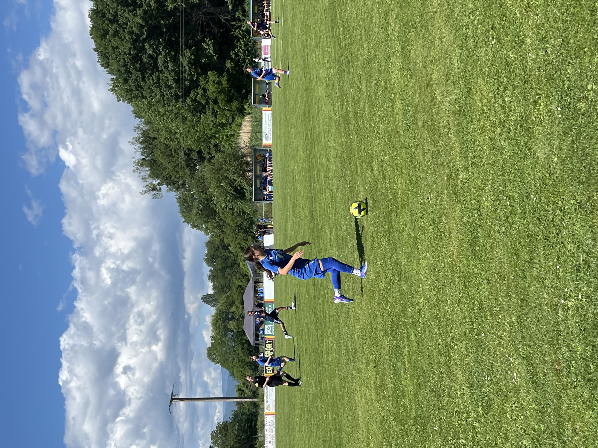 An outdoor daytime scene depicts a women's soccer match in progress on a green grass field in Porte du Ried, France. The sky is bright blue with numerous large, white cumulus clouds.
In the foreground, slightly right of center, a female soccer player wearing a royal blue uniform (jersey, shorts, socks, white and purple cleated shoes) is captured in mid-stride, running from right to left across the frame. Her dark hair is tied back in a ponytail. A yellow and black soccer ball rests on the grass slightly ahead of her right foot.
In the mid-ground, from left to right:
* A person in a black referee uniform is running.
* Two other players in royal blue uniforms are running.
* A player in a dark uniform is also running.
* Along the perimeter of the field, several advertising banners are visible, including one displaying "SPORT" and a "CA" logo, and another with French text "L'AUBERGE AU PONT DE LA PECHE."
* Behind the banners, two dark-colored pop-up tents are erected.
* Further right, two yellow and black open-sided dugouts or shelters contain seated individuals.
The background consists of a dense line of green deciduous trees bordering the field. Beyond the trees, distant blue-grey mountains are faintly visible on the horizon. A tall wooden utility pole with electrical lines is present on the far left side of the image, extending into the sky. The grass field shows small scattered white flowers, likely daisies. Strong sunlight illuminates the scene.