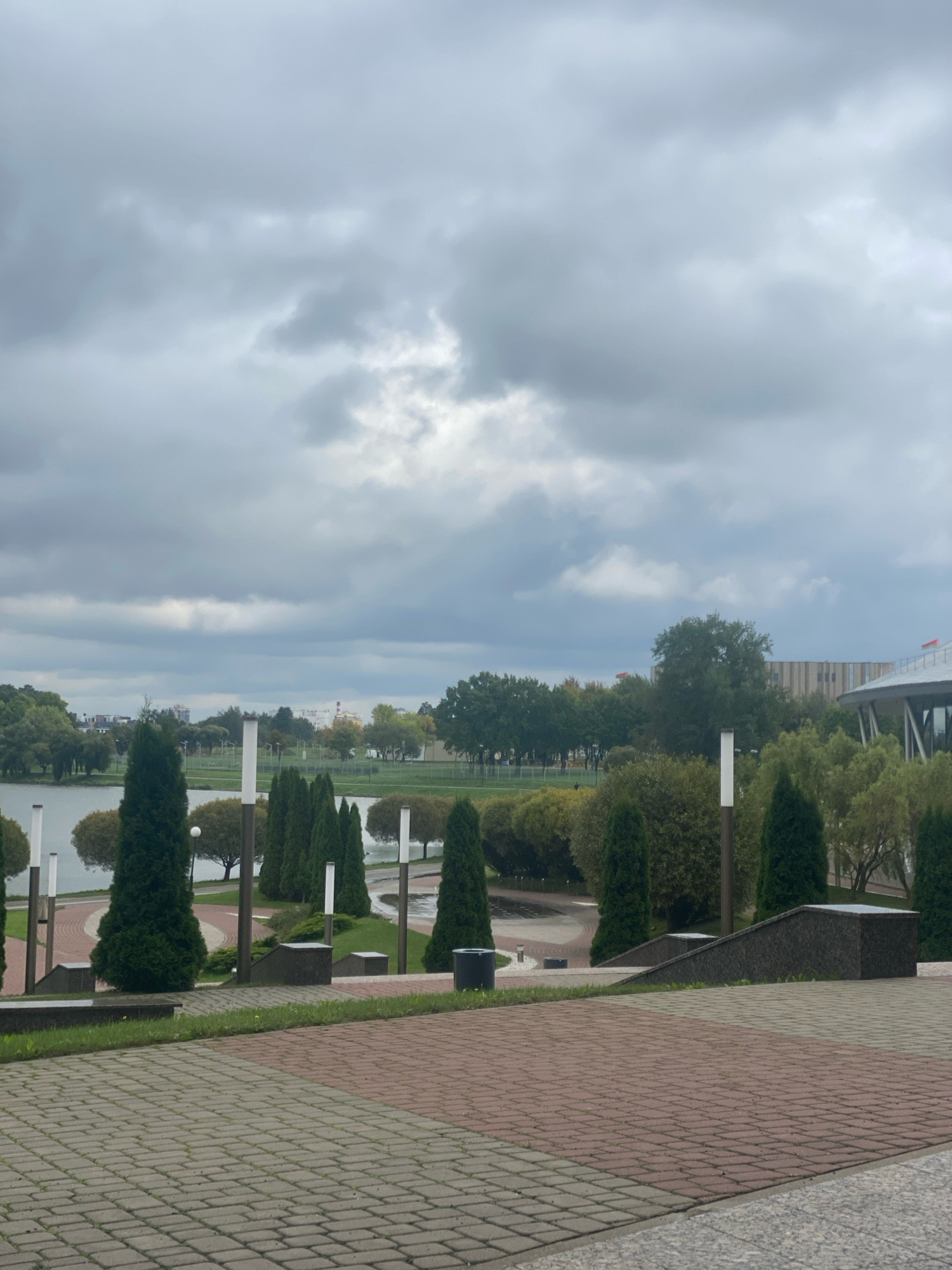 This image depicts a park area in Minsk, Belarus, likely near a body of water such as a lake or river, given the presence of a shoreline and reflections. The sky is overcast with heavy, gray clouds, suggesting a potentially cool or damp day. The foreground features a paved walkway made of red and gray interlocking bricks, leading up a slight incline. Beyond the walkway, a grassy area is visible with several dark, rectangular planters or benches.
Further into the park, a collection of tall, slender evergreen trees, likely cypresses or similar varieties, are arranged in a line. These trees are interspersed with modern, white-lighted lampposts. A winding paved path can be seen curving through the park, bordered by more greenery and some rounded bushes. A body of water is visible in the middle ground, with trees and buildings lining its opposite bank. The buildings in the background appear to be a mix of modern structures, possibly office buildings or a stadium, with some featuring striped facades and curved roofs. No people are visible in the image. The overall impression is of a well-maintained urban park designed for recreation and aesthetic appeal, despite the somber sky.