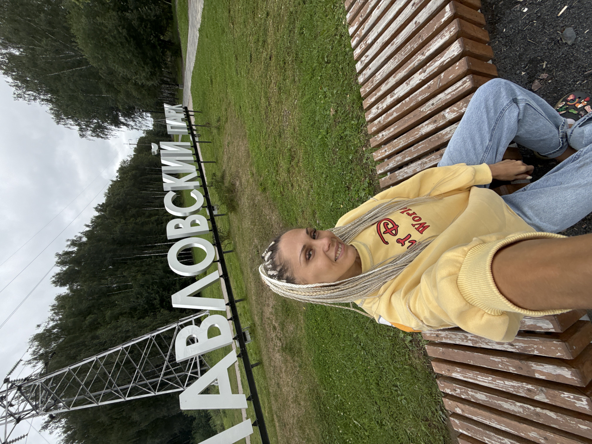 In Rusanovo, Russia, a woman with blonde braided hair smiles at the camera while sitting on a wooden bench in a park. She is wearing a yellow sweatshirt with a red Disney logo and light blue jeans. Behind her, large white letters spell out "ЗАВЛОВСКИЙ ПАРК" (Zavlovsky Park), indicating the location. A tall power line tower stands to the left of the park sign, and lush green trees form a backdrop. The ground is covered with grass and a paved walkway is visible to the right. The sky is overcast.