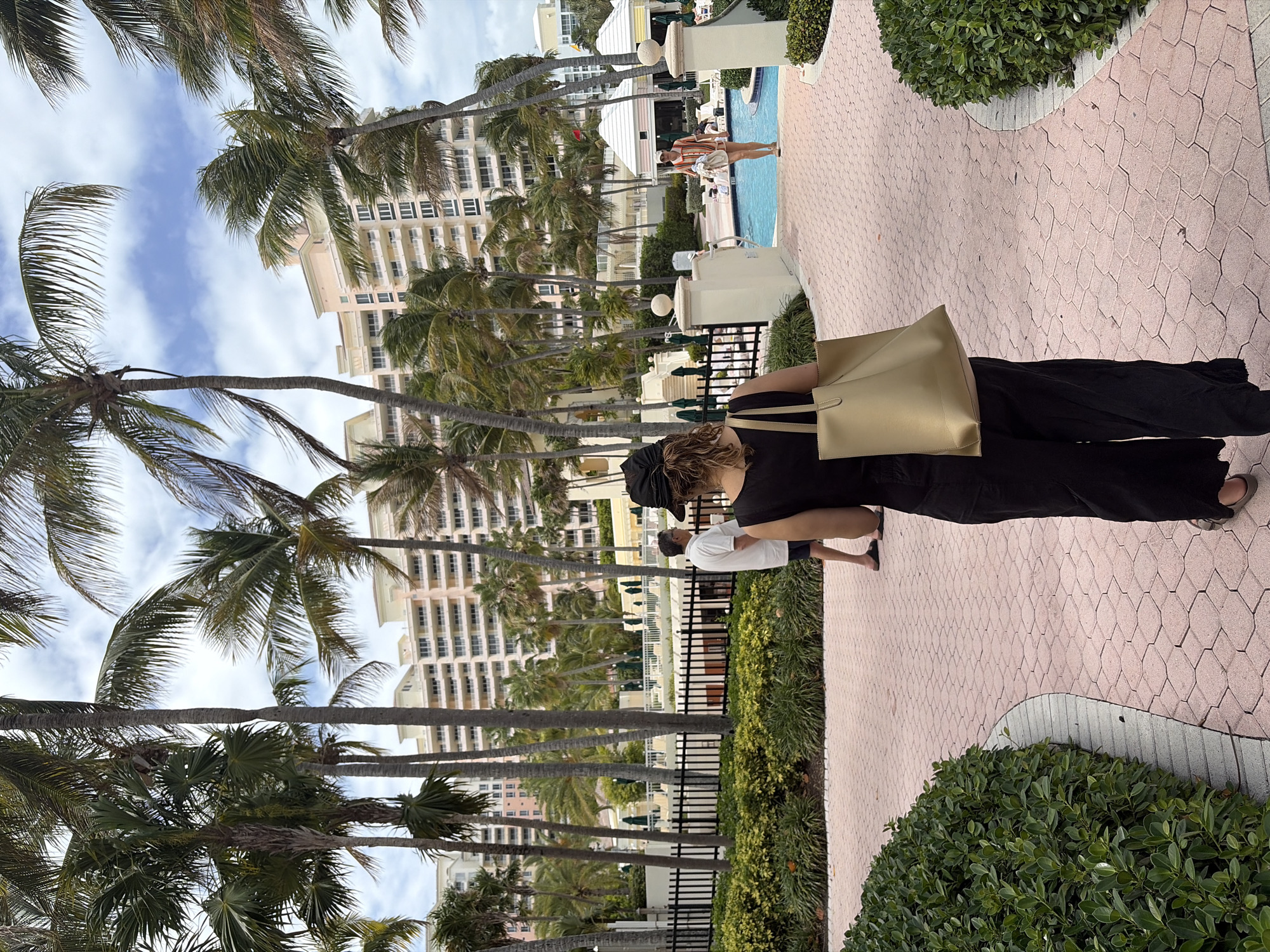The image depicts an outdoor scene in Key Biscayne, United States. A walkway paved with pink, geometric-patterned tiles is bordered by green bushes. Palm trees line the left side of the walkway, partially obscuring a tall, light-colored building in the background. A black metal fence separates the walkway from the palm trees and the building.
Two people are walking on the pathway, partially facing away from the camera. One person is wearing black clothing and carrying a large, light-colored tote bag. The other person is wearing a white shirt. In the background, a swimming pool area is visible with people near the water. The sky is partly cloudy.