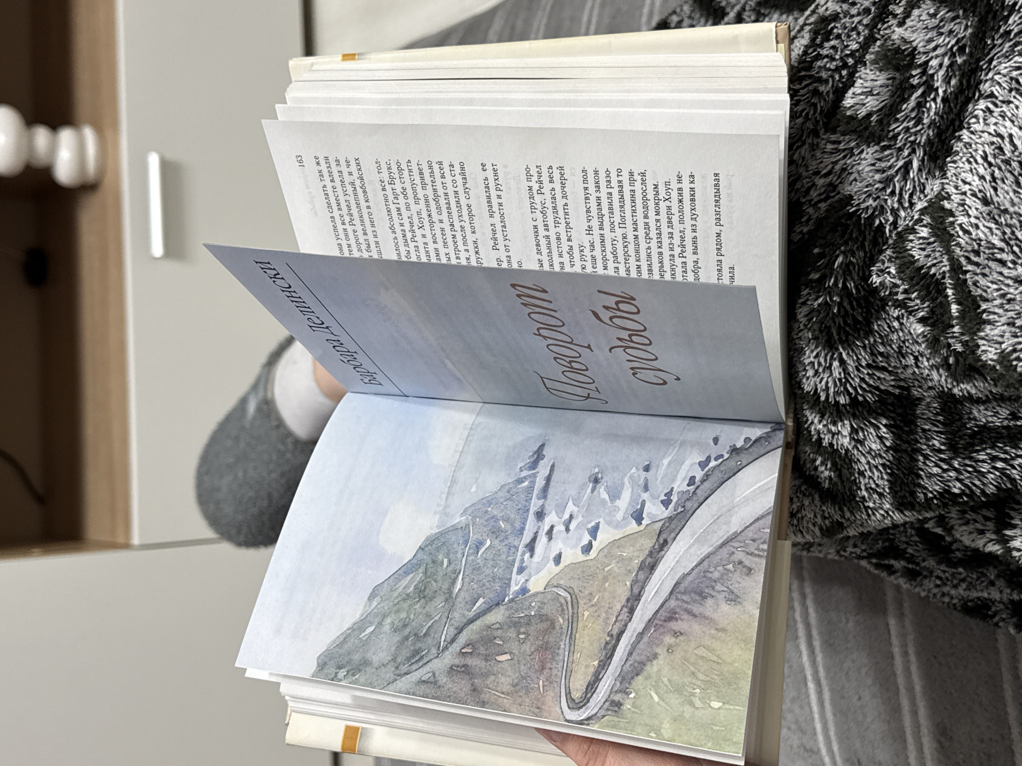 Here is a description of the image:
A book is held open, displaying a landscape painting on the left page and text in Russian on the right. The left page features an illustration of a mountain road winding through a valley, rendered in watercolor with muted blues, greens, and grays. The right page is filled with lines of Russian text, with some words highlighted in a darker ink. The book is being held by a person with a gray sleeve in the background. The right side of the frame is covered with a black and gray fuzzy blanket.