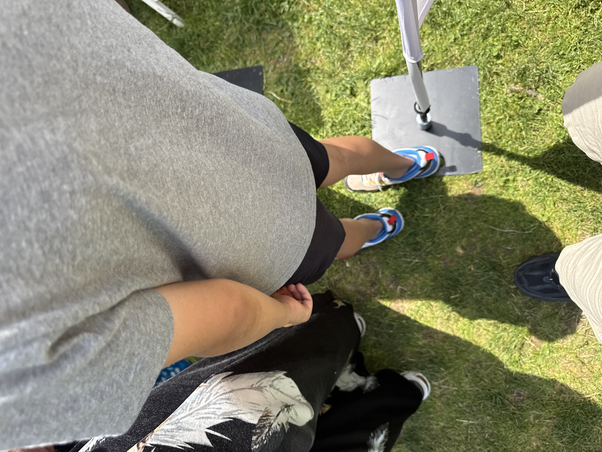 An overhead, sunlit view depicts three individuals standing on a grassy outdoor surface in Sofia, Bulgaria. A central person, wearing a grey t-shirt, black shorts, and blue-and-white athletic shoes, stands with their right foot on a dark grey square base that supports a vertical pole. The left foot is on the grass. To the left, a second person is partially visible, wearing black clothing with a white floral or feather pattern and a white-soled shoe. At the bottom of the frame, the lower body of a third person, wearing light-colored pleated trousers and black shoes, is visible. Direct sunlight casts distinct shadows across the green grass.