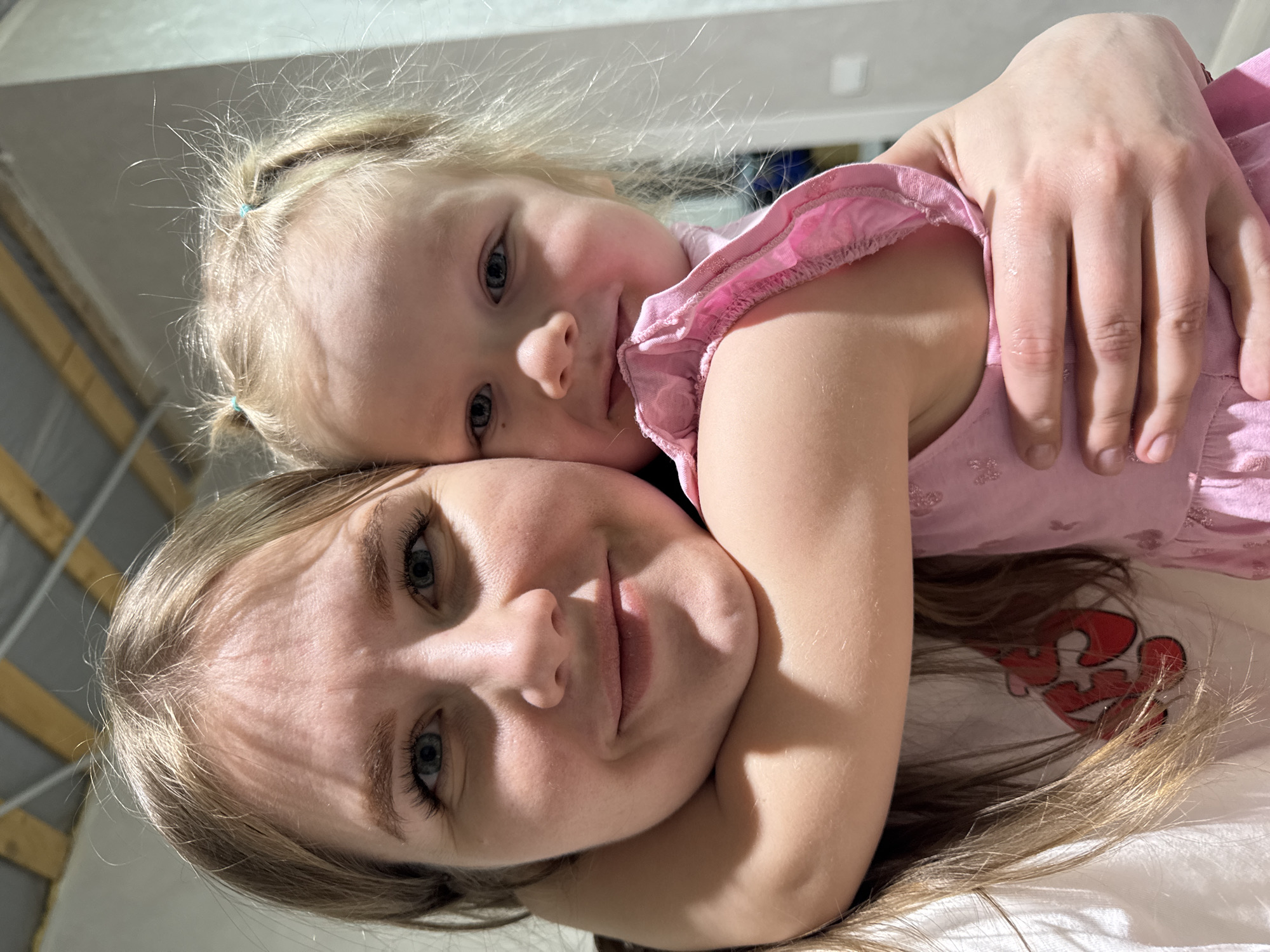 An adult female and a young female child are captured in a close-up, front-facing image taken indoors in Gornyi (Dvurechenskoe S/P), Russia. The adult female, positioned on the left, has blonde hair, light skin, and blue eyes, and is smiling towards the camera. She wears a white shirt, partially revealing a red graphic on the chest area. The child, positioned on the right and slightly behind the adult, also has blonde hair styled in two small pigtails secured with blue elastics, light skin, and blue eyes, and is smiling towards the camera. She wears a light pink dress with ruffled shoulder straps and subtle patterned details. The child's left arm is draped around the adult female's neck and right shoulder, indicating an embrace or close proximity. The background is blurred, showing a light-colored wall section on the right and a ceiling with visible wooden beams and a lighter panel overhead.