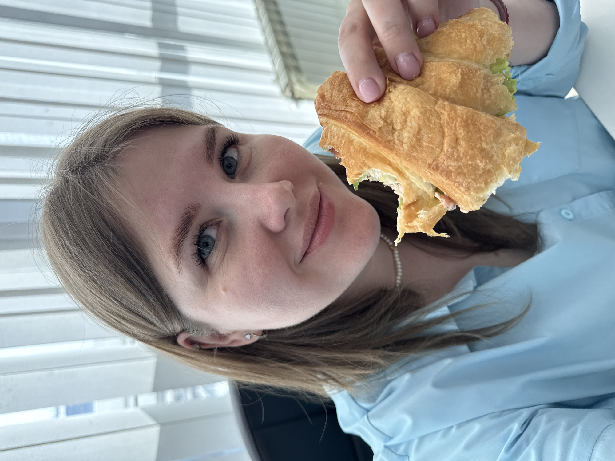 A young woman with blonde hair and blue eyes is holding a partially eaten sandwich in her right hand. She is wearing a light blue collared shirt and a pearl necklace. The background includes venetian blinds and what appears to be a dark-colored object, possibly a computer monitor. The location is assumed to be Perm, Russia.