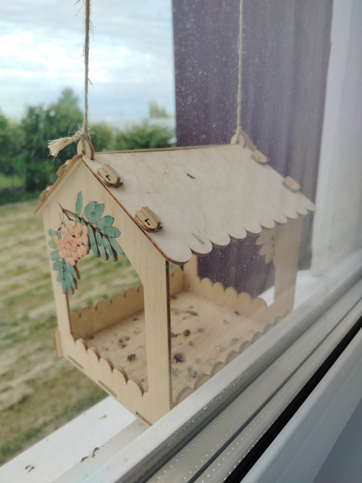 A light-toned wooden bird feeder, configured as a miniature house with scalloped roof and base edges, is suspended by a frayed natural fiber rope outside a window in the city of Смалявічы, Belarus. A decorative element featuring a branch with light orange berries and teal leaves is present on one side of the feeder's roof, which is visibly assembled with small wooden interlocking tabs. The base of the feeder contains numerous dark specks, indicative of bird droppings and seed debris. The perspective is through a windowpane, which displays visible dust and water spots, particularly on the right side. Beyond the window, an overcast sky is visible above a blurred background comprising green trees and a pale green field. A dark, out-of-focus structure is discernible in the far right portion of the background.