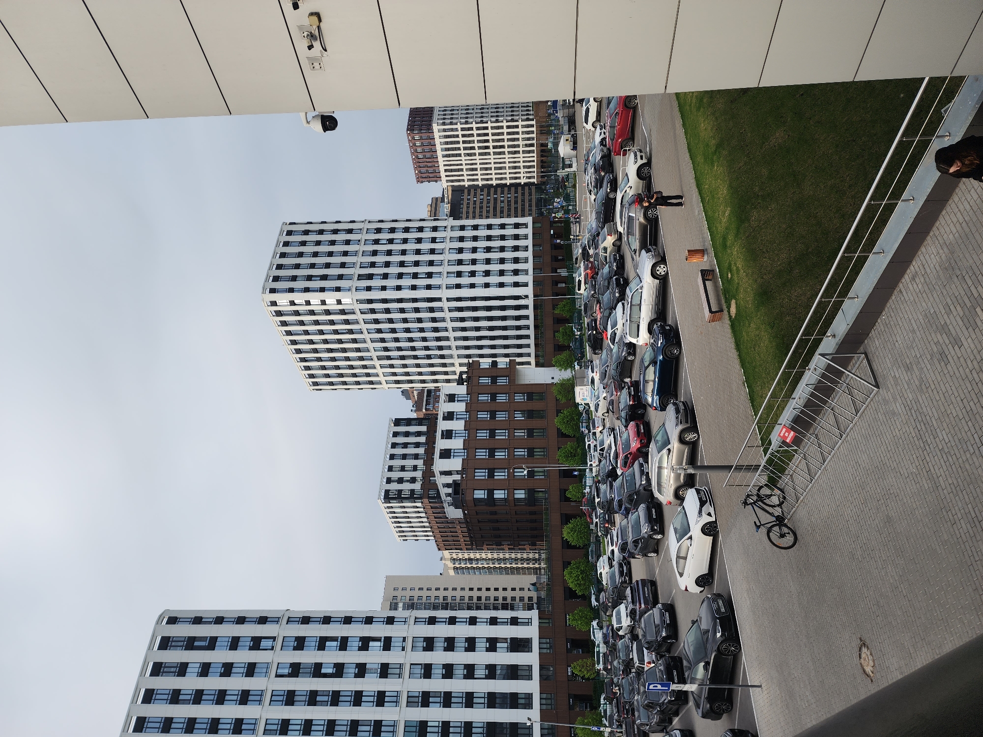 The image shows a densely packed parking lot adjacent to several modern, multi-story buildings, likely residential apartments, in Kazan, Russia. The buildings are characterized by white or brown facades with repeating rectangular window patterns.
A long line of cars of various colors (black, white, red, silver, and blue) stretches along the parking lot. There are people standing around some of the cars. Next to the parking lot is a grassy area.
To the right of the parking lot is a paved area with a metal railing. A black bicycle is parked on the pavement. There is a person standing near the railing.
In the upper part of the image, the edge of a building is visible. Two surveillance cameras are mounted on the building. The sky is overcast.