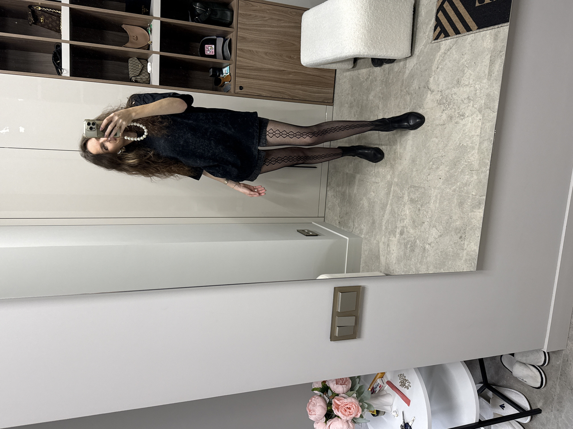A person takes a photograph of themself in front of a mirror. The individual is wearing a black t-shirt, patterned tights, and black ankle boots. A string of pearls is visible around their neck. The background includes storage cabinets, a white stool, and part of a rug. Below the mirror is a white round table with floral decorations. The setting appears to be indoors.