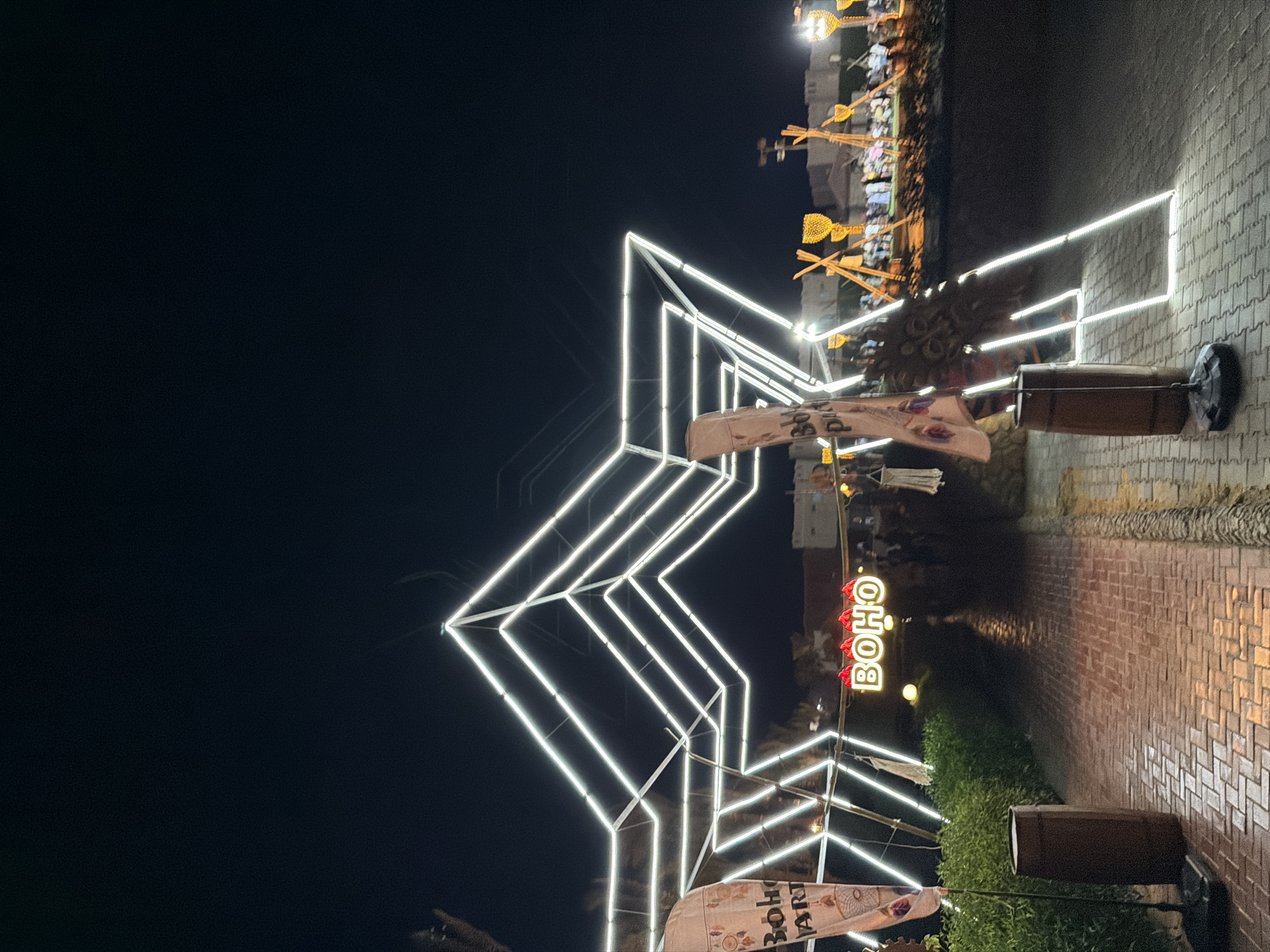 The image shows a nighttime scene in Sharm El Sheikh, Egypt. A large, geometric structure outlined in white neon lights dominates the foreground. To the left, a flag with the text "BOHO ART" is visible. In the center, a neon sign spells out "BOHO". The ground in front of the structure is paved with bricks, and there are bushes and barrel-like objects on either side of the walkway. In the background, a crowd of people can be seen gathered, with more illuminated structures and lights visible, suggesting a festival or outdoor event. The sky is dark, indicating it is nighttime.