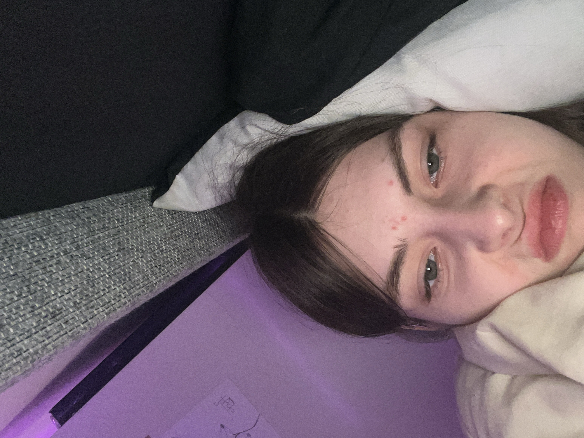 A close-up, slightly skewed, selfie-style photograph depicts a young woman lying down. Her face is the primary subject, revealing fair skin, dark hair, and a few blemishes on her forehead. Her eyes are open and appear to be a greenish-gray. She is resting her head on a white pillow or fabric. The background includes a section of what appears to be a gray couch and a dark, possibly black, blanket or clothing item. The lower portion of the image shows a purple-lit wall. There is no evidence to suggest that this photo was taken in Saint Petersburg, Russia.