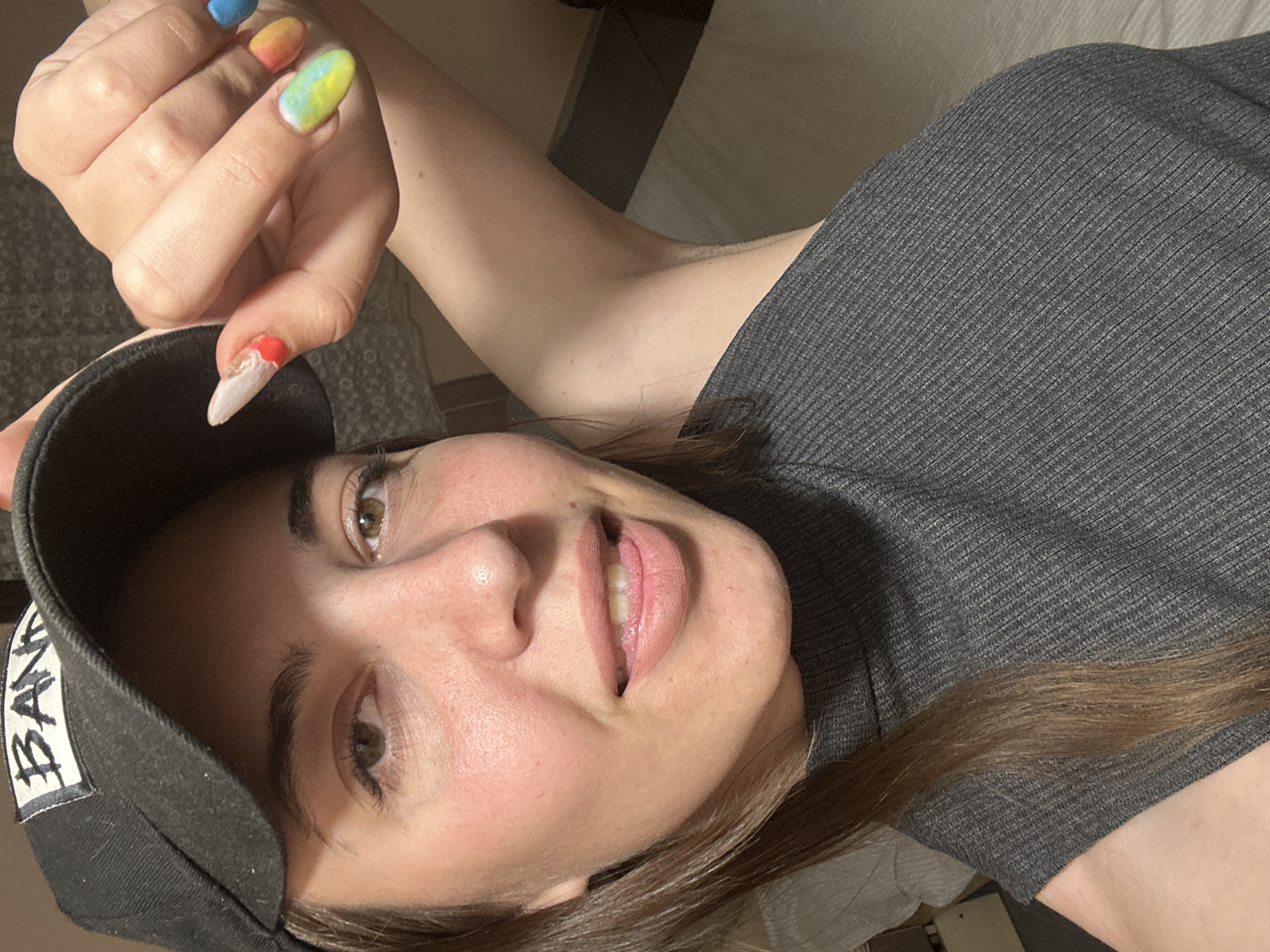 A young woman is captured in a close-up, tilted portrait, seemingly lying down and looking towards the viewer. She has light brown hair, brown eyes, and fair skin, with a faint blush on her cheeks and a subtle smile. Her lips are a natural pink and slightly parted, revealing her bottom teeth.
She is wearing a dark grey, ribbed short-sleeved top. A black baseball cap is worn backward on her head, with a white label visible on the side that reads "BAND" in a stylized font.
Her left hand is raised towards her face, showcasing her long, almond-shaped nails, each painted with a distinct vibrant design. The nails, from what is visible, include one in solid blue, another with an orange-to-yellow gradient, a third with a yellow-to-blue gradient, and her thumb nail which is white with a red tip at the base.
The background is indistinct and appears to be an indoor setting with soft, ambient lighting. The overall mood of the image is casual and relaxed. The location context provided is Sirakovo, Bulgaria.