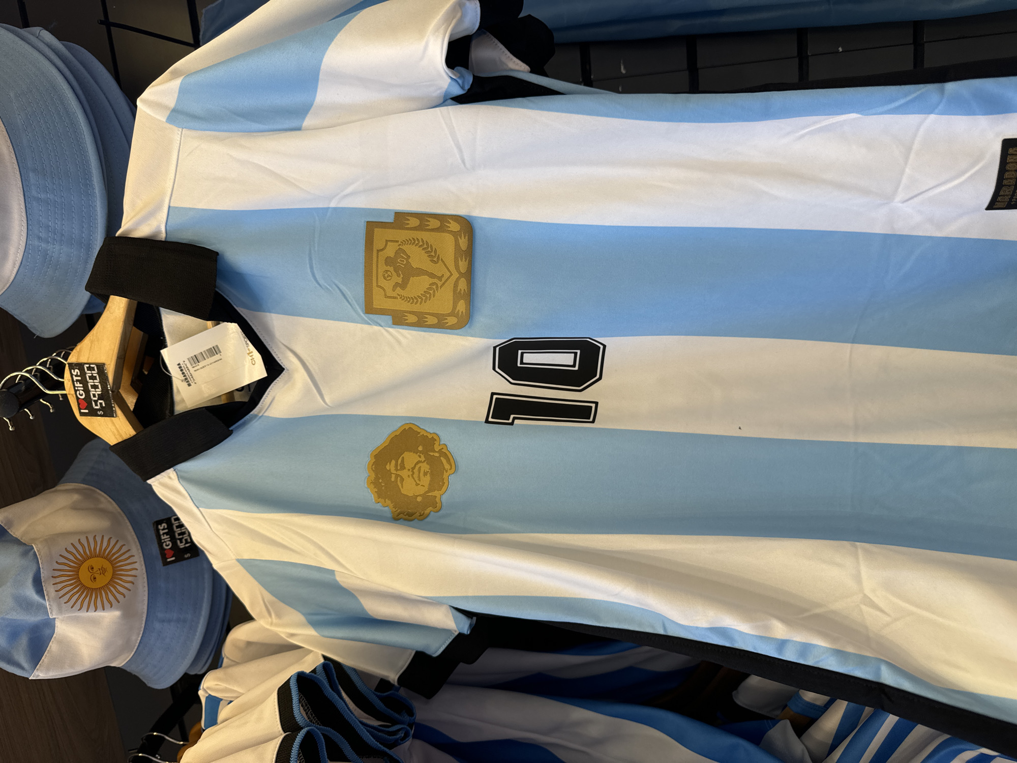 The image shows a striped light blue and white Argentina soccer jersey hanging on a rack in a store. The number 10 is visible in black print on the jersey, along with gold patches showing a soccer player and a portrait of Diego Maradona. Also on the rack is an Argentina-themed bucket hat and clothing tag with a price in Argentinian pesos. The location is likely a souvenir store in Buenos Aires, Argentina, given the presence of national team merchandise and currency on the tags.