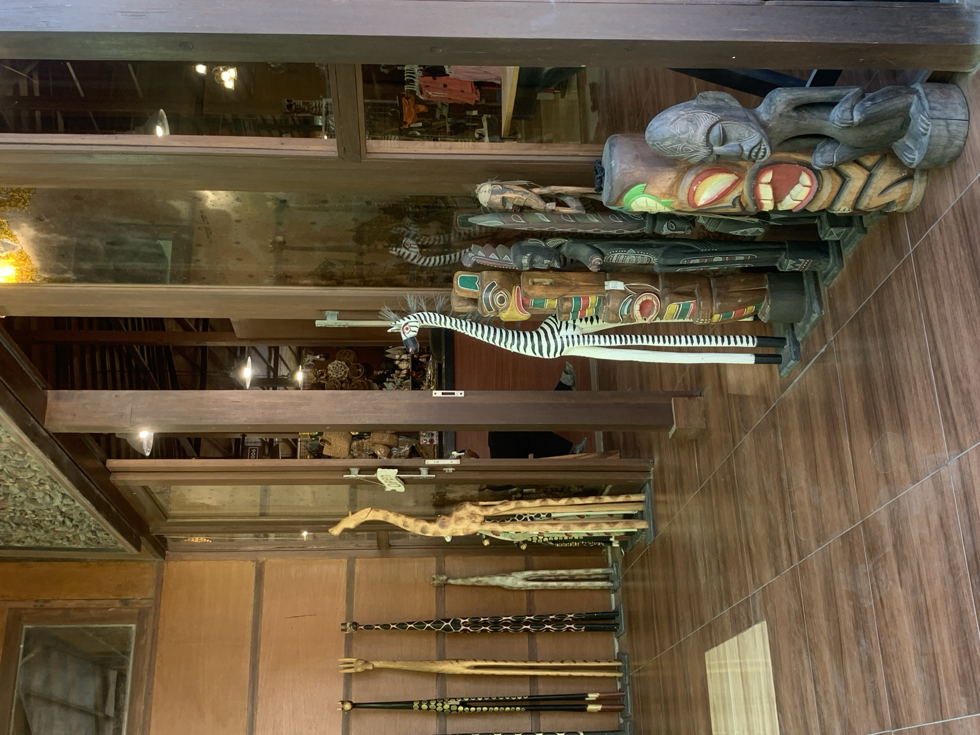 The image shows the interior of a shop in North Kuta, Indonesia, filled with wooden carvings. In the foreground, a collection of stylized animal and tribal figures are displayed. To the left, several tall, slender wooden giraffes, varying in design and height, stand against a wood-paneled wall. One giraffe is intricately carved and painted with a spotted pattern, while others are more simplistic. Further to the left, three dark, elongated figures with pointed tops are partially visible.
Dominating the center of the frame is a large, striped wooden zebra figure standing tall. Next to it, a series of carved wooden totems with distinctive facial features and colorful patterns are arranged in a line. These totems include figures with large mouths, eyes, and intricate carvings. To the right of the totems, a dark wooden carving of a seated figure, possibly human, is placed on a circular base.
The shop's interior features wooden floors with a tiled pattern and exposed wooden structural beams. Large glass doors and windows reveal the interior of the shop, with racks of clothing visible in the background. A sign indicating "Open" is attached to a glass door. The lighting appears to be from overhead fixtures, creating a warm ambiance. The overall scene suggests a souvenir shop or a gallery specializing in wooden crafts.