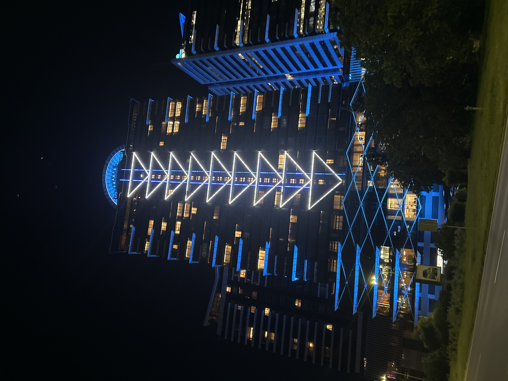 This is a night-time photograph of the Capital Fort office building in Sofia, Bulgaria. The building is illuminated with a prominent triangular light display that runs down its facade, interspersed with blue lights. To the right of Capital Fort, another modern office building is visible, also lit up with blue accents. In the foreground, a dark road curves to the left, with a grassy median and some trees. The sky above is dark, with a few faint celestial objects visible.