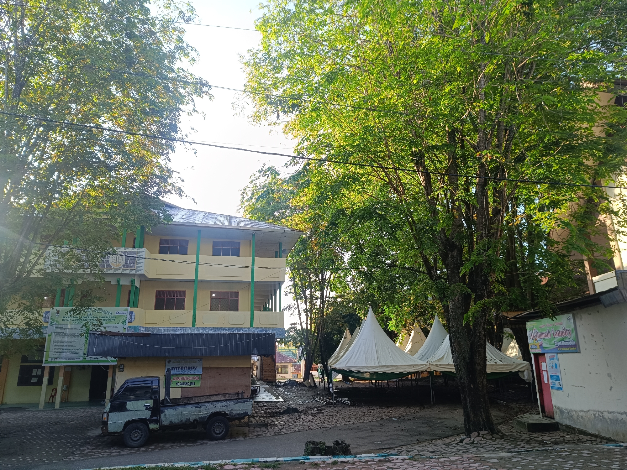 The image depicts an outdoor scene in Lhokseumawe, Indonesia, featuring a three-story yellow and green building with a sign indicating it is the "SEKRETARIAT PENERIMAAN SANTRI BARU DAYAH ULUMUDDIN KOTA LHOKSEUMAWE" (New Student Admission Secretariat for Dayah Ulumuddin Lhokseumawe City). In front of the building, there is a weathered pickup truck parked. To the right, there are several white tents erected, suggesting a temporary event or activity. Adjacent to the tents is a small building with a sign for "Ummah Laundry" and a "FOTOCOPY" sign suggesting a photocopying service. The environment includes lush green trees casting shadows on the ground and overhanging power lines. The pavement is composed of textured, patterned stones.