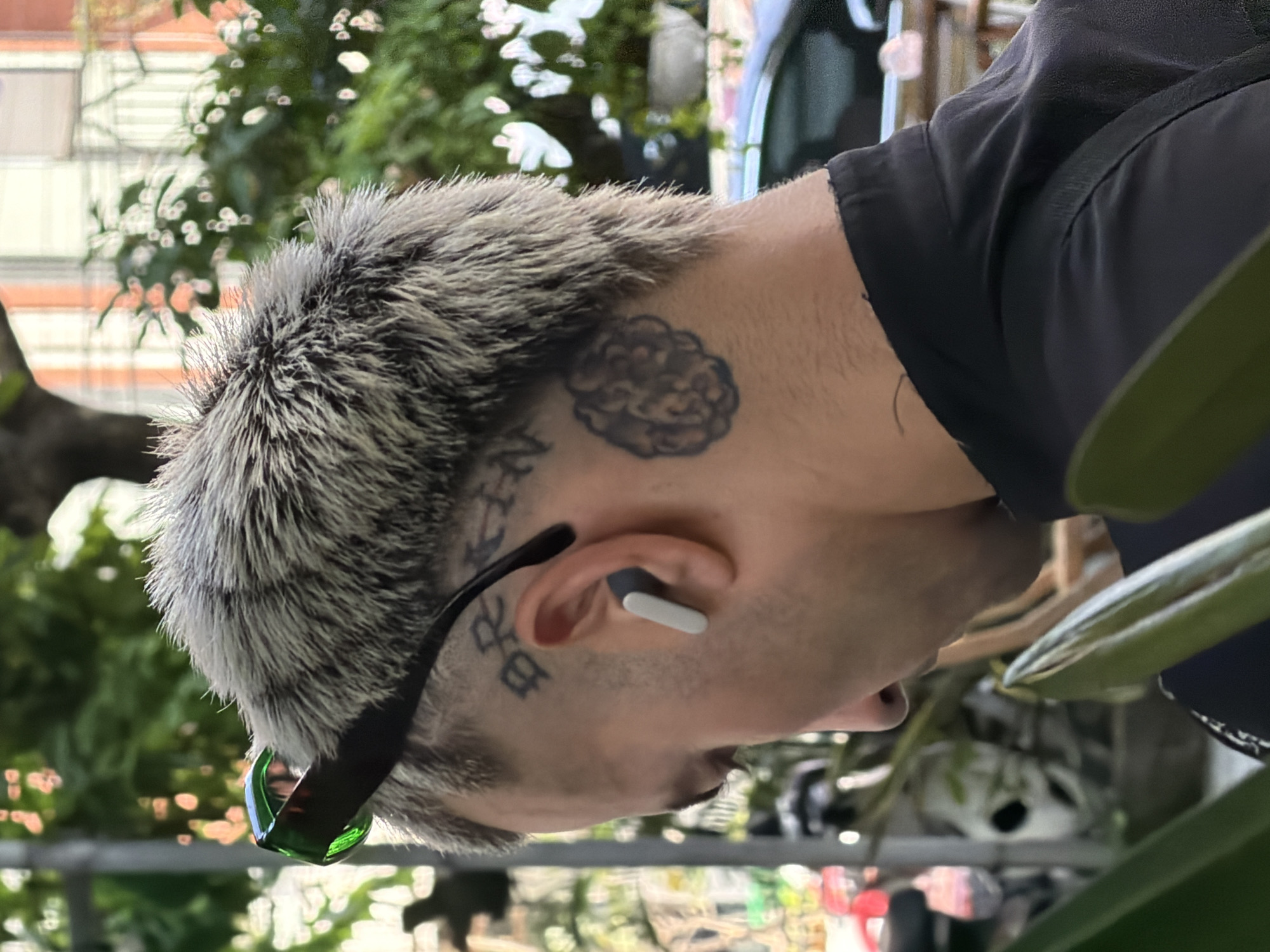 A close-up, slightly rotated image focuses on a person's head and upper back. The person has a grey, short-spiked hairstyle. Tattoos are visible on the person's head: on the upper-left side of the head is a brain-shaped tattoo, a design of Vietnamese characters behind the ear, and more Vietnamese characters near the left temple. The person wears sunglasses with black frames and green tinted lenses, and a white and grey wireless earbud is visible in the left ear. The person is wearing a black shirt. A blurred background shows greenery and what appears to be an urban cityscape, possibly in Đà Nẵng, Vietnam.