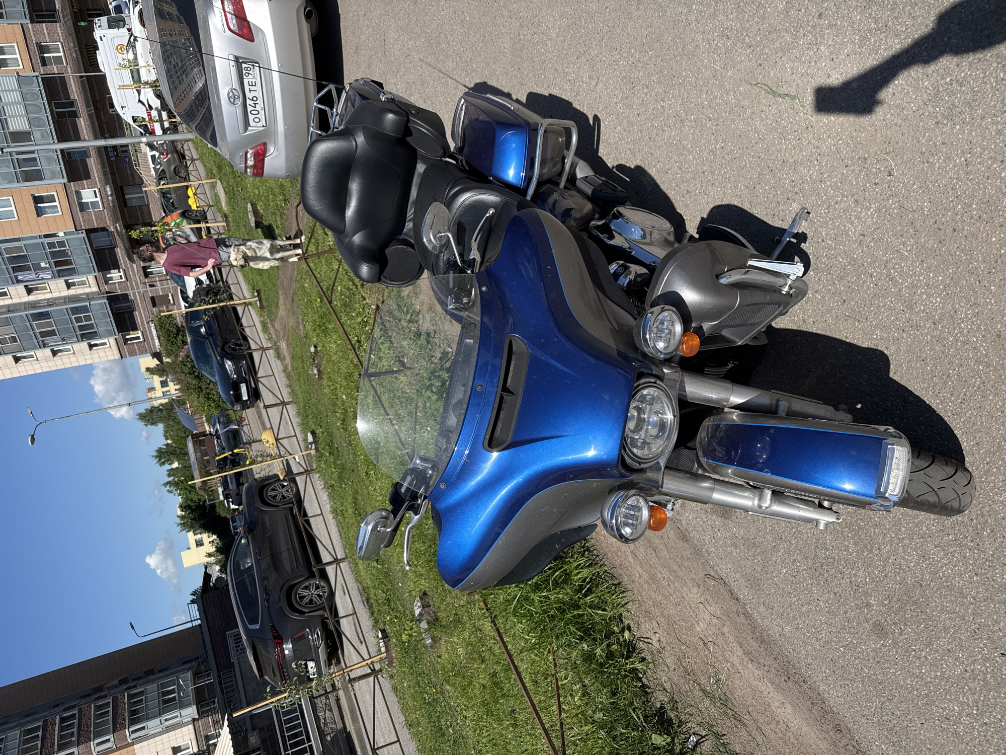 This media file captures an outdoor street scene in Sinyavino, Russia, under bright daylight with a clear blue sky and scattered white clouds. The perspective is slightly elevated, looking across a paved road and grassy verge towards residential buildings.
The main subject is a prominent blue and grey touring motorcycle, parked in the foreground, partially on the asphalt and partially on a grassy strip. It features a large windshield, chrome handlebars, multiple headlights, and plush black leather seats. Distinct shadows indicate a sunny, likely midday or early afternoon.
In the mid-ground, a person wearing a burgundy t-shirt and patterned shorts walks a light-colored dog on a leash along the grassy area. Behind them, a silver Toyota Camry sedan is parked, displaying the license plate "О046ТЕ 98 RUS". Further down the street, a black SUV and other vehicles are also parked.
The background consists of multi-story residential buildings, likely apartment blocks, with visible windows and balconies. The scene depicts a calm, everyday urban or suburban environment.