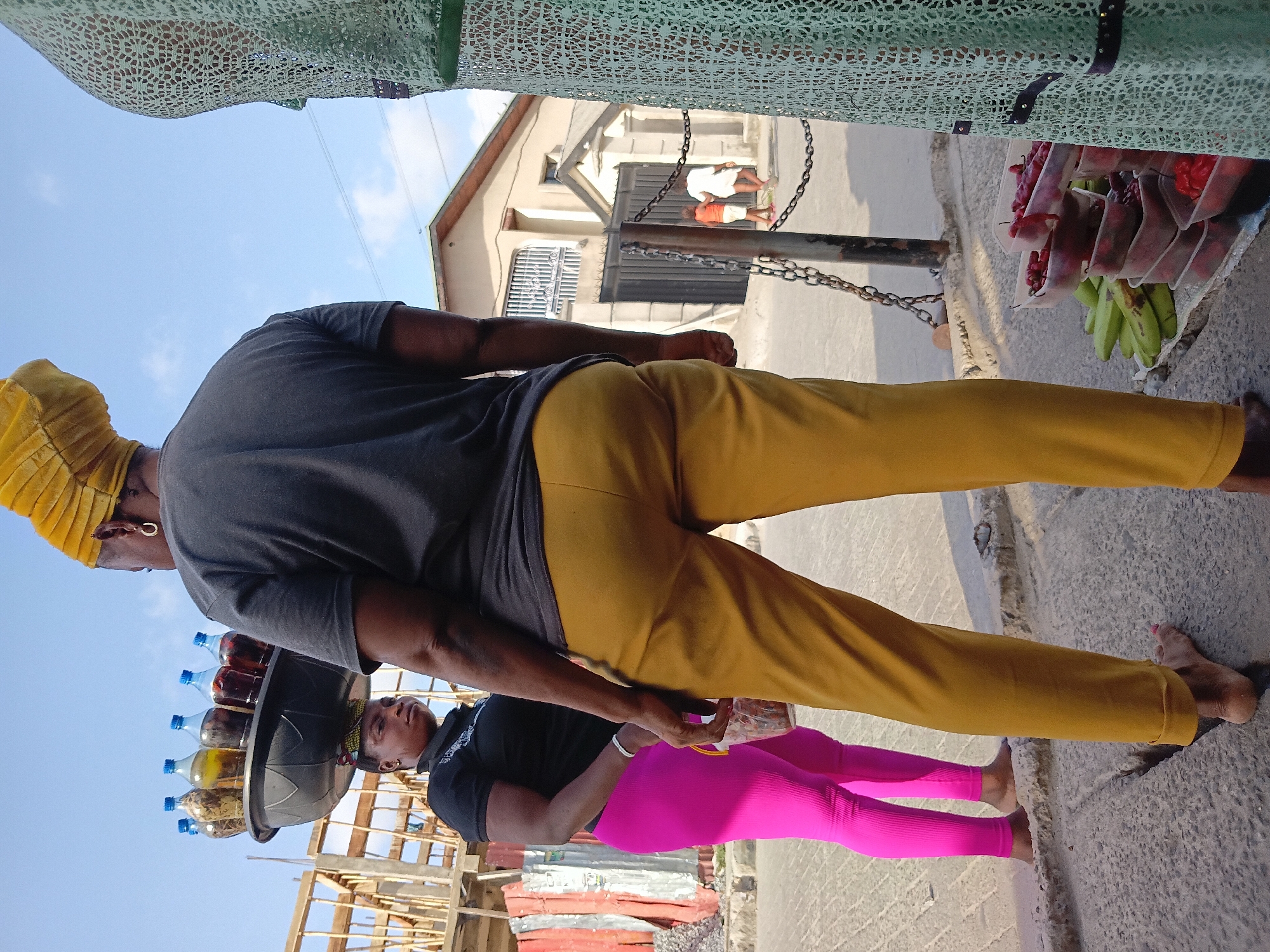A ground-level medium shot taken in Ado, Nigeria, shows two women in the foreground on a paved street. The woman nearest the camera is wearing a dark grey shirt and mustard-yellow pants, with a gold-colored headwrap. She is walking to the right and away from the viewer, barefoot. The second woman is walking in the same direction, wearing a black shirt and bright pink leggings. She is also barefoot and holding a clear plastic bag. On the head of the first woman is a large, shallow black bowl holding clear plastic bottles filled with varying shades of liquid.
To the right of the frame, visible behind the first woman's legs, are stacked bananas and red fruits contained in clear plastic containers. These items are partially obscured by a lacy green fabric hanging from above.
Further back, slightly left of center, are a white building with dark metal doors and two individuals. One is wearing white, and the other orange.
The ground is concrete, and the background features blue skies and glimpses of construction. The image is taken in daylight, and shadows are visible on the ground.