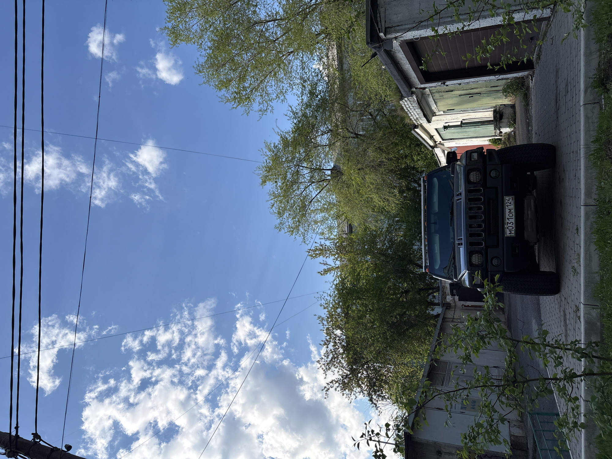 The image shows a black Hummer H2 parked on a paved street in what appears to be an older, possibly residential area. The car's license plate, M031OM 124, indicates it is registered in the Krasnoyarsk region of Russia. The background consists of weathered garage structures with varied door styles and paint colors, suggesting a slightly neglected or older urban environment. Overhead, power lines stretch across a partly cloudy sky with trees visible on both sides of the street. The lighting appears to be natural daylight. There are no people visible in the scene.