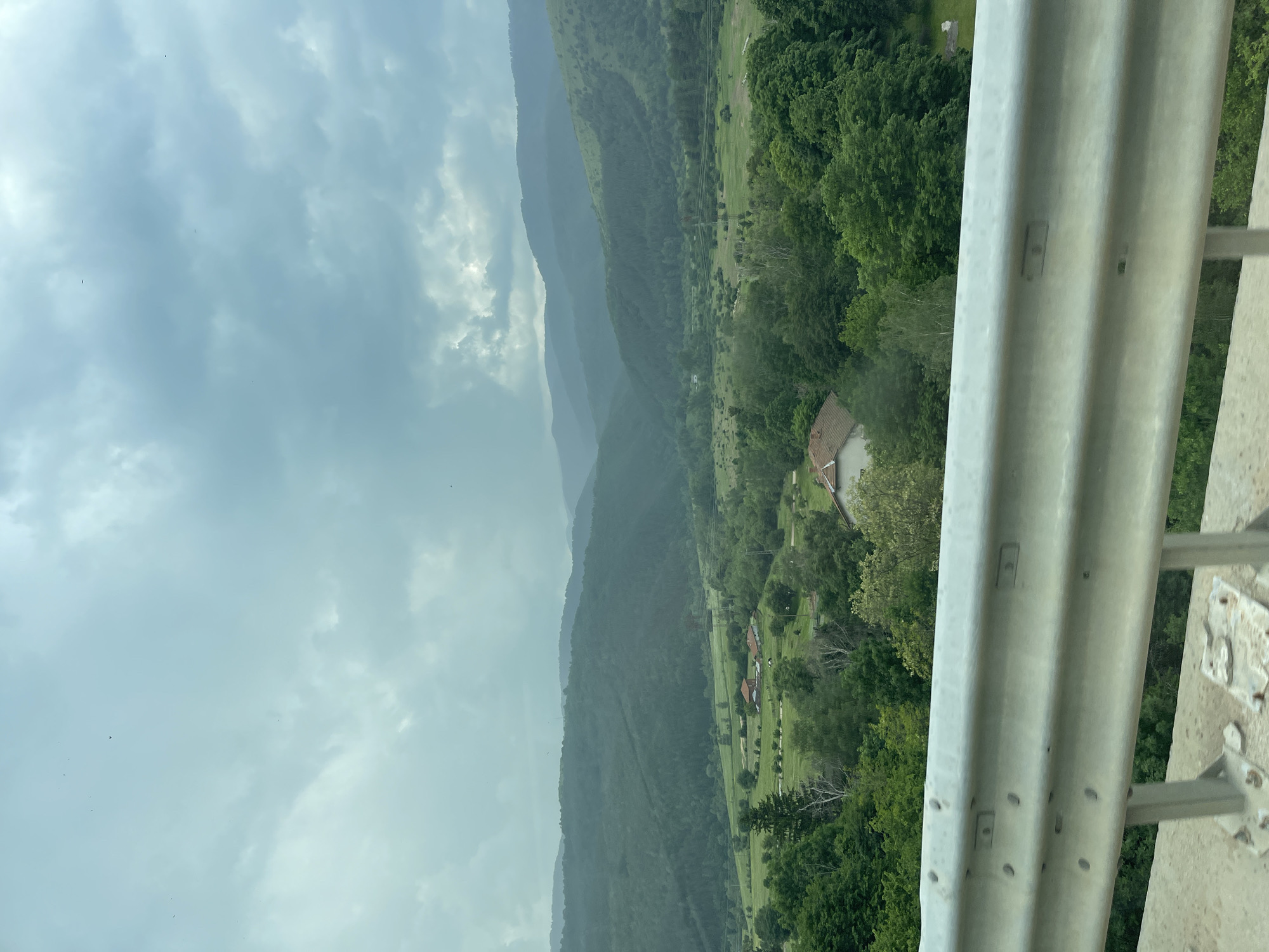 This detailed description pertains to an image captured in the city of Anton, Bulgaria.
The scene presents a panoramic view of a mountainous and heavily forested landscape under an overcast sky, observed from an elevated perspective, likely a bridge or roadside.
In the immediate foreground, a metallic grey guardrail occupies the lower approximately one-fifth of the frame. The guardrail consists of two horizontally corrugated sections, secured by multiple visible bolts and supported by vertical posts. Below the guardrail, a concrete base or road surface is visible, along with some metal plates and debris on the ground. Green foliage, comprising trees and bushes, is visible behind and below the guardrail, extending down an embankment.
The midground reveals a verdant valley interspersed with agricultural fields and clusters of trees. Several residential structures with reddish-brown tiled roofs are present within this valley, indicating human settlement. One prominent house is situated slightly right of center, with another to its left. A partial view of a third house is visible in the bottom right corner, just behind the guardrail. A power line tower (pylon) with visible wires stands in the midground, slightly right of center, spanning across the valley.
The background is dominated by successive layers of rolling hills and mountains, densely covered with dark green forest. These landforms recede into the distance, exhibiting a gradual atmospheric haze that lightens their appearance further away.
The sky, occupying the upper portion of the image, is heavily overcast with varying shades of grey and light blue, suggesting cloudy or potentially inclement weather. Small, dark, indistinct specks, likely birds or insects, are scattered across the sky, particularly in the upper left and central-right regions.
No human figures or explicit actions are discernible within the image. The overall environment is rural and natural, characterized by extensive forestry and limited human infrastructure in the valley.