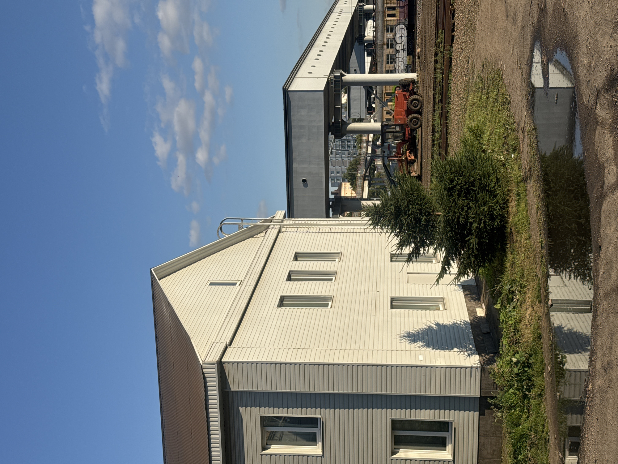 The image depicts an outdoor scene on a sunny day with blue skies and scattered clouds, likely in Khimki, Russia. In the foreground, a large puddle reflects a white building with a brown roof, which is partially visible on the left side of the frame. This building, constructed with horizontal siding, features several windows. Adjacent to the building, a small evergreen tree casts a distinct shadow on its facade.
The midground reveals a gravelly area with railroad tracks. A bright orange construction vehicle, possibly a grader, is parked on the tracks. Beyond the vehicle, a long, elevated concrete structure supported by pillars stretches across the frame. This structure appears to be part of an overpass or a similar elevated transportation or infrastructure element. In the background, several multi-story buildings are visible, suggesting an urban or suburban setting. The overall impression is one of an industrial or transport-related area with ongoing construction or maintenance.