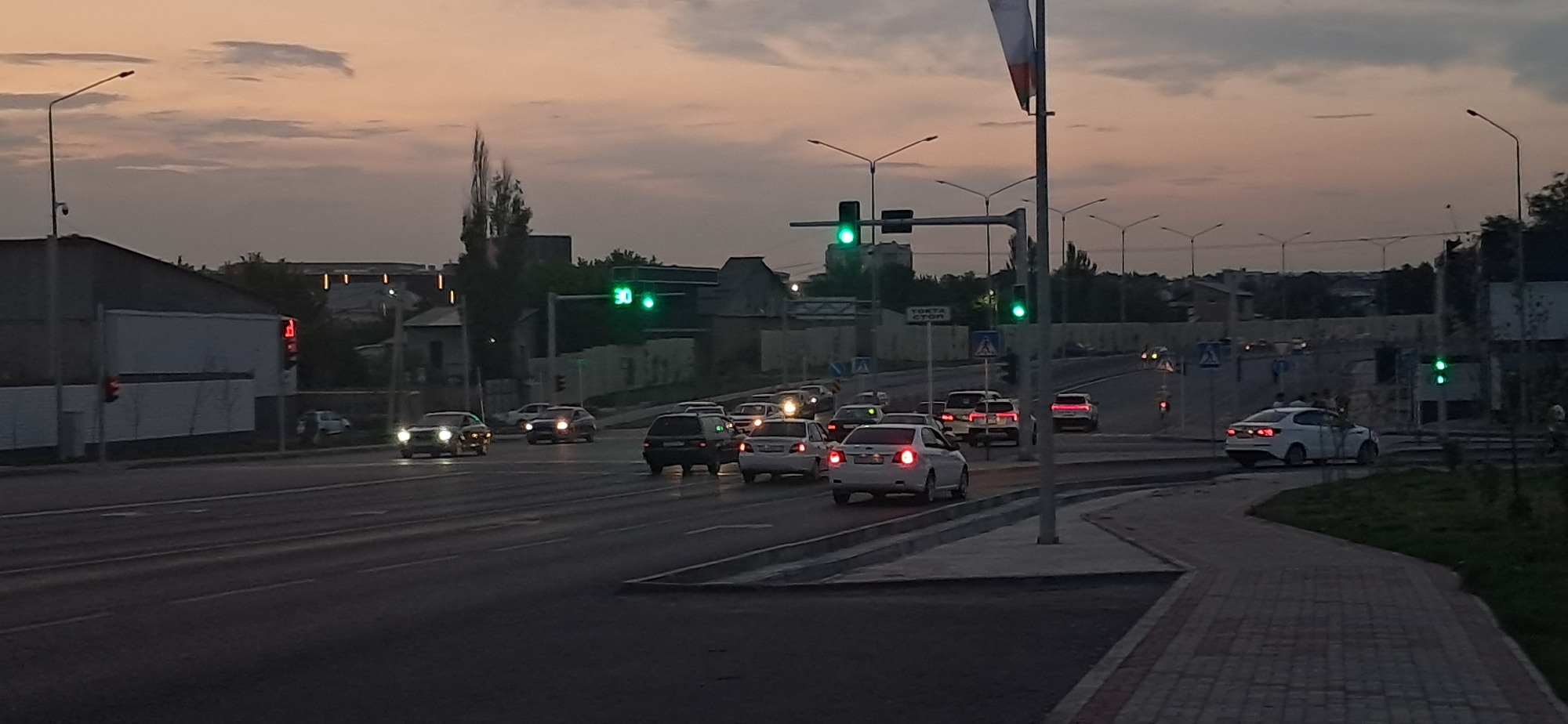 The image depicts a street intersection at dusk in an urban setting. Several cars are visible on the road, some of which appear to be stopped at a traffic light. The traffic lights themselves are illuminated, with at least one showing a green light. Streetlights are also visible, casting a dim glow on the scene. Buildings line the side of the road, and trees and other vegetation can be seen in the background. The sky is overcast with hues of orange and gray, indicating the transition from day to night. A pedestrian walkway with a brick pattern is visible in the foreground. There are no people clearly visible in the scene.
