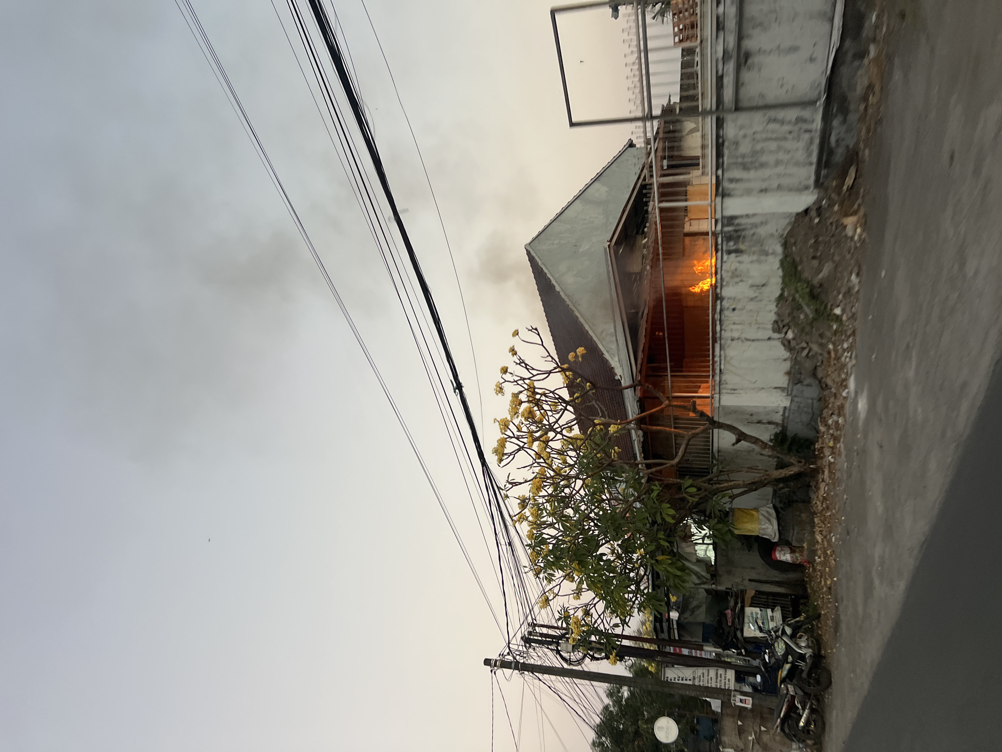 The image depicts a residential house fire in Denpasar, Indonesia, as evidenced by the dense smoke billowing from the roof and the visible flames on the ground floor. The fire appears to be actively burning, with orange flames prominently displayed. Several motorcycles are parked in front of the property, suggesting a residential area. A tree with yellow flowers stands to the left of the house, partially obscuring the view of the fire. Overhead, a complex network of electrical wires spans across the sky, common in urban and suburban Indonesian settings. The overall scene is one of emergency and destruction.