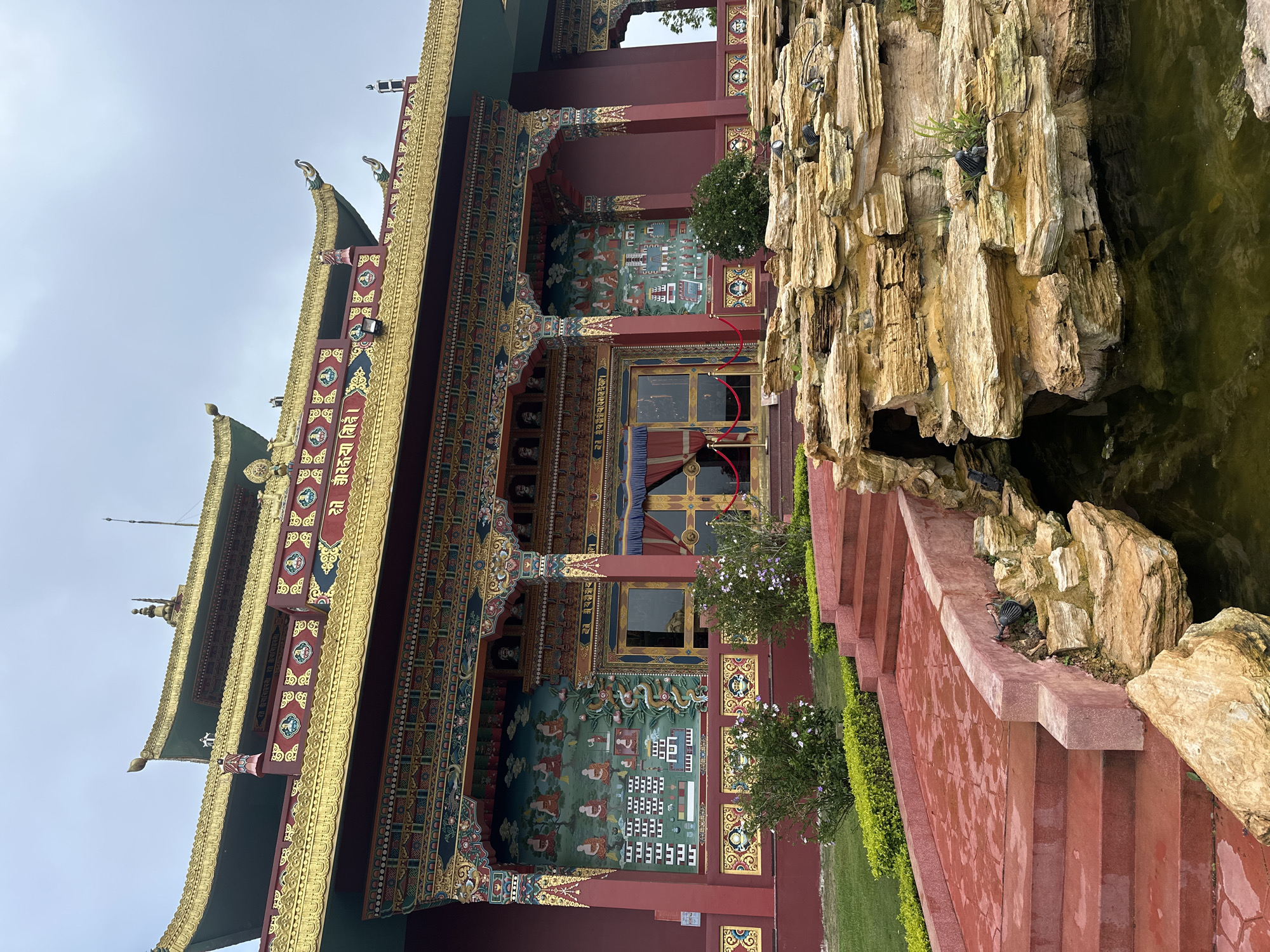A Buddhist temple with vibrant red walls and ornate golden roofs stands under a cloudy sky. The building features intricate murals depicting scenes of Buddhist life and ornate golden decorative elements. Red steps lead up to the entrance, bordered by green bushes and flowering plants. In the foreground, a pond with rocky formations is visible, with a small waterfall or stream feature. The temple is located in a peaceful, well-maintained garden setting.
