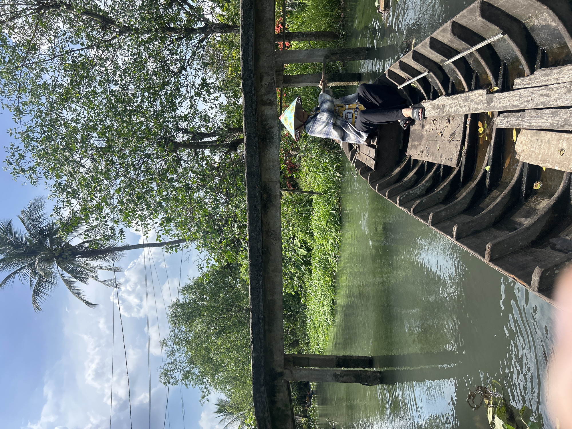 A person wearing a conical hat and a plaid shirt is seated in a wooden boat on a narrow waterway. The boat is moving, indicated by the wake. The person is holding a wooden pole, likely used for steering or propulsion. The waterway is surrounded by lush green vegetation, including trees and reeds. A concrete bridge with two supporting pillars spans the waterway in the background. Power lines are visible in the upper left corner of the image. The sky is blue with scattered white clouds. The scene appears to be in a rural area, possibly a canal or river in An Hữu, Vietnam, given the vegetation and the presence of a boat as a mode of transportation.