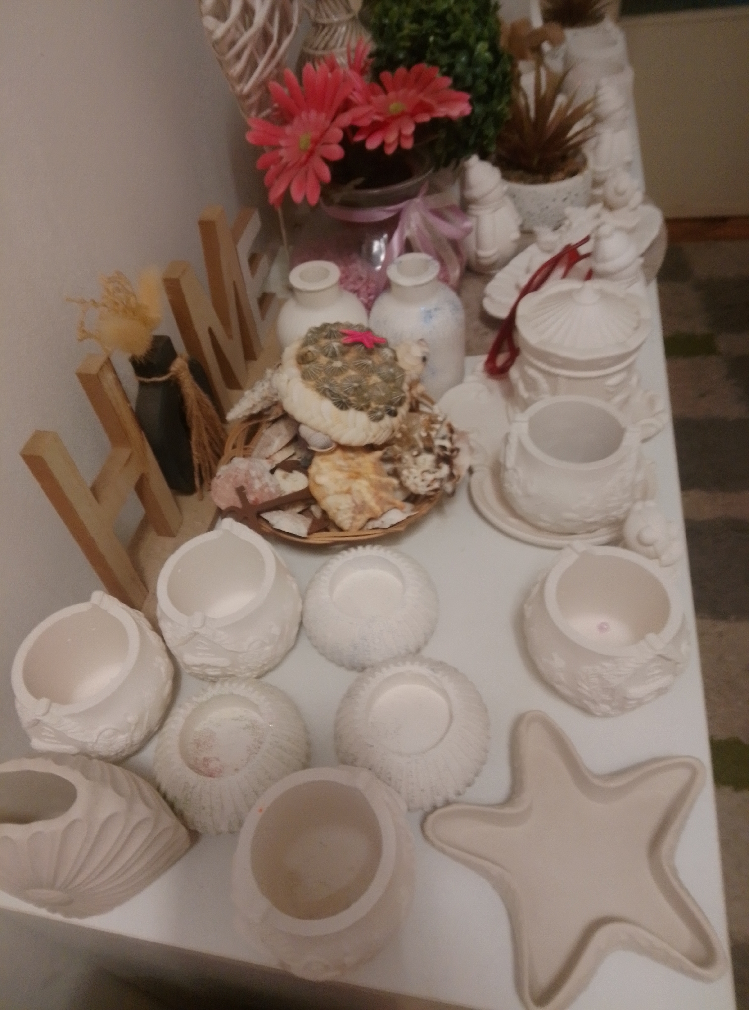 The image shows a collection of white ceramic decorative items arranged on a white surface, likely a shelf or table. The items include various shaped pots, containers, and a starfish-shaped dish. Some of the ceramic items are decorated with embossed patterns. In the background, there are some artificial pink flowers and greenery, as well as wooden letters spelling out "HOME". A wicker basket filled with shells and rocks is also visible. The overall scene appears to be a display of decorative crafts or home decor items. The location in the city of Wedel, Germany, is not discernable from the image content.