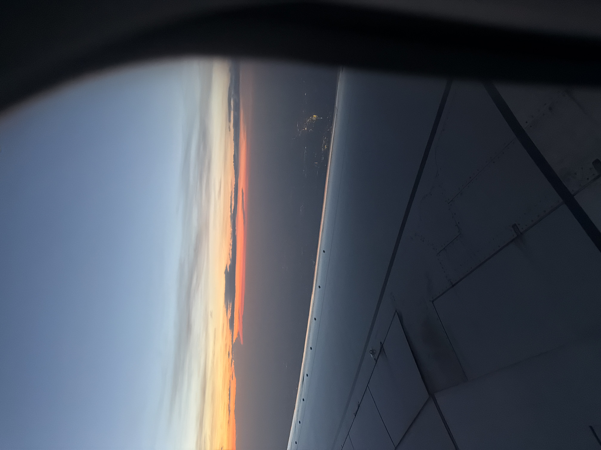 A view from an airplane window shows the wing of the plane and a sunset or sunrise. The sky transitions from blue to orange, suggesting twilight. Below the horizon, faint lights indicate a distant cityscape. The location is unlikely to be Перевозное, Russia, as it's a small village with minimal nighttime illumination.