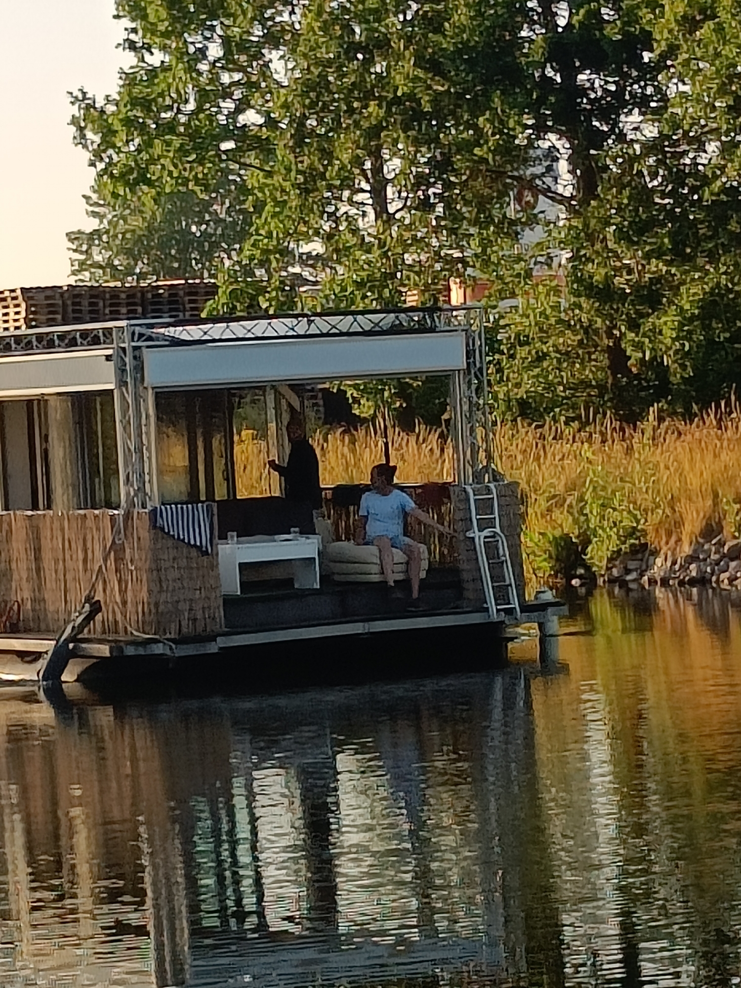 In Haacht, Belgium, a houseboat is docked on a body of water. On the deck of the houseboat, two individuals are present. One person, dressed in a black garment, stands near the entrance of the houseboat. Another person, wearing a light blue t-shirt and shorts, is seated on a cushioned bench to the right. A white table and some striped cushions are also visible on the deck. The houseboat has a modern structure with a metallic frame supporting a roofed area. The surrounding environment features lush green trees and tall, dry reeds along the water's edge. The water reflects the trees and the houseboat, creating a rippled pattern.