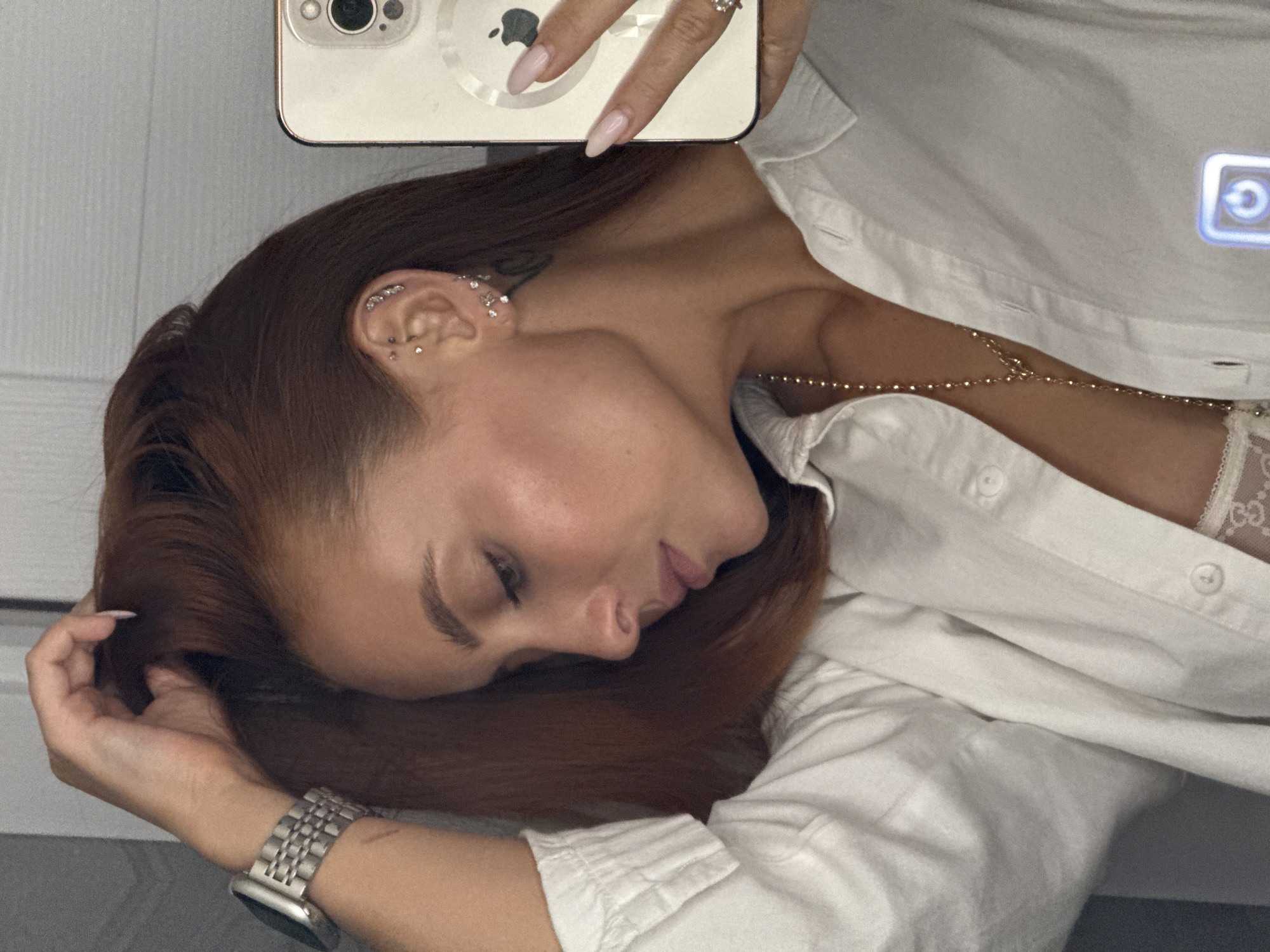 A medium-shot image captures a woman lying down, seemingly taking a selfie. The woman has auburn hair, fair skin, and is wearing a white button-down shirt. A gold necklace is visible at her neckline. Her left ear is adorned with multiple piercings. She is holding a smartphone with a white case, and a watch is visible on her left wrist. The background appears to be a simple, neutral-toned interior. The location in the city of Chaikovsky, Russia, cannot be determined from this image.