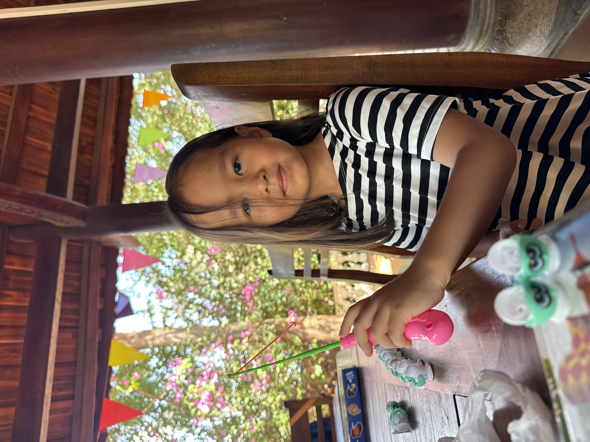 A young girl sits at a wooden table playing a fishing game. She is wearing a black and white striped shirt. In her right hand, she holds a pink plastic fishing rod with a green line and orange hook. On the table are several small white and green toy fish, some marked with the number "3," along with the game box. A white plastic bag is also visible on the table.
The scene appears to be in an open-air structure with wooden beams and supports. In the background, outside the structure, there are trees and flowering bushes, and colorful pennant flags are strung up. Based on the visual elements, the location appears to be in a casual outdoor setting, possibly a resort or restaurant in Mũi Né, Vietnam, considering the typical architecture and tropical vegetation of the area.