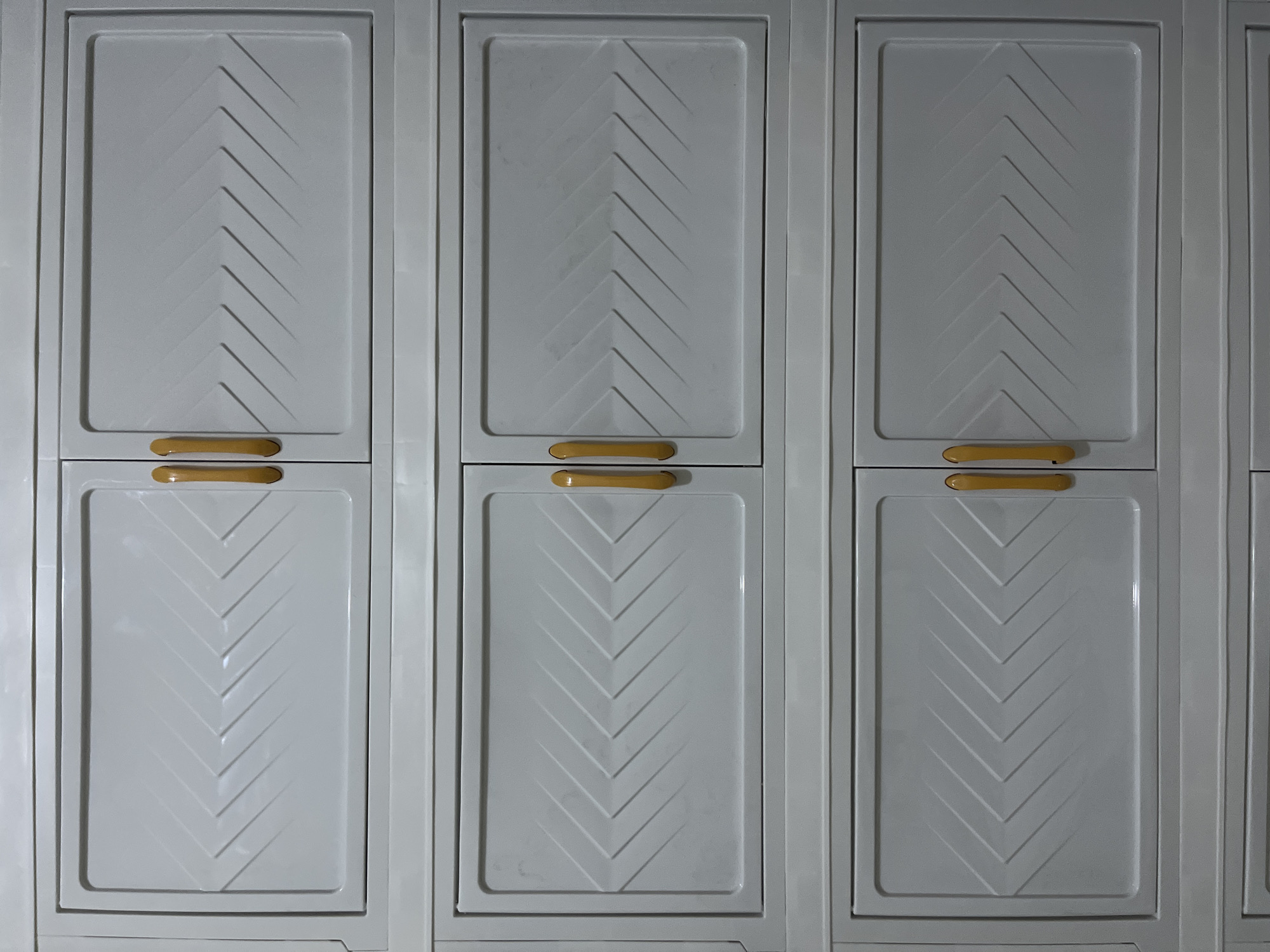 The image shows a close-up of a light gray modular cabinet with multiple doors. Each door has a rectangular recessed panel with a chevron pattern embossed into it. The handles are a glossy yellow-brown color and are located in the center of the right edge of the left-side doors and the left edge of the right-side doors, indicating that each section has two doors. There are three visible rows of these double doors, with a total of six doors visible in full. The cabinets appear to be made of plastic. The background is a plain, dark wall. There are no people or other objects visible in the image. The image does not provide any specific location within East Jakarta, Indonesia.
