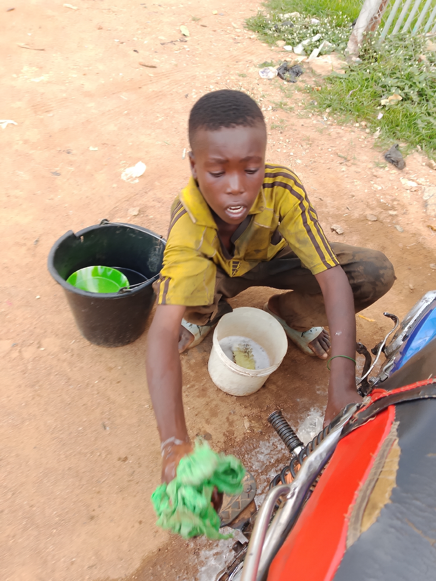 A young boy is washing a motorcycle on a dirt ground. He is wearing a yellow t-shirt with black stripes on the sleeves and dark pants. He is kneeling down and holding a green cloth in his right hand. In front of him, there are two buckets: a black one with a green bowl inside and a white one with soapy water. The background shows dry dirt, grass, and some scattered trash. A metal fence is visible on the right side of the image. The scene appears to be an outdoor area in a potentially unpaved part of a city.