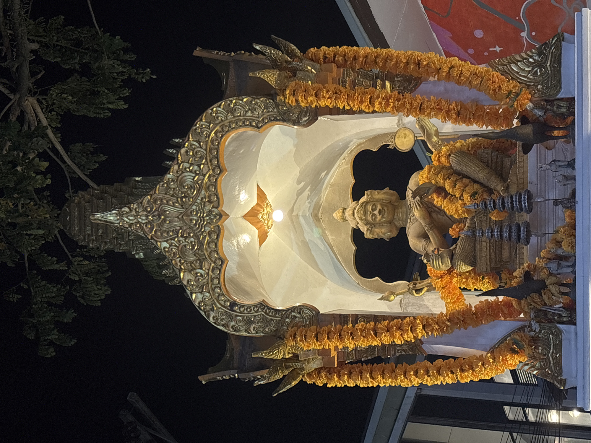 Here's a description of the image:
The image shows a richly decorated shrine, likely located in Choeng Thale, Thailand. The shrine is adorned with gold-colored ornamentation, floral garlands (likely marigolds), and a central figure of a deity. The deity is also decorated with floral garlands. The shrine is illuminated from within, with the light emphasizing its decorative details. The background reveals a combination of natural elements (trees, sky) and architectural features, suggesting an urban setting. The lighting implies it is either nighttime or a dimly lit indoor location.