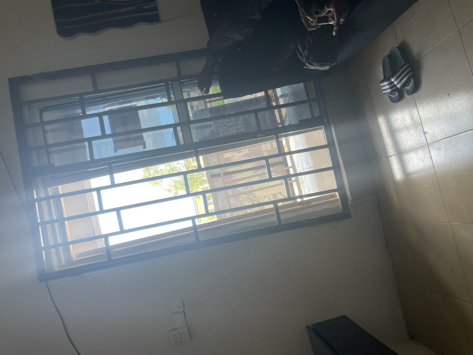 The image depicts an interior scene of a room with a large window covered by a metal grate. Sunlight streams through the window, creating bright highlights and casting shadows on the tiled floor. On the floor, near the bottom right of the frame, a pair of black Adidas slides with white stripes are placed. To the right of the slides, a dark couch is visible, with a person's legs and feet resting on it. The person is holding a white video game controller in their right hand.
The walls are a light color, and a black furniture item is partially visible on the far left. The window grate is made of black metal bars and appears to be a security feature. Through the grate, an outdoor scene can be observed, including some greenery and the edge of a building. The overall impression is of a simple, lived-in space. The specific location in Jalingo, Nigeria is not directly evident from the visual cues within the image itself.
