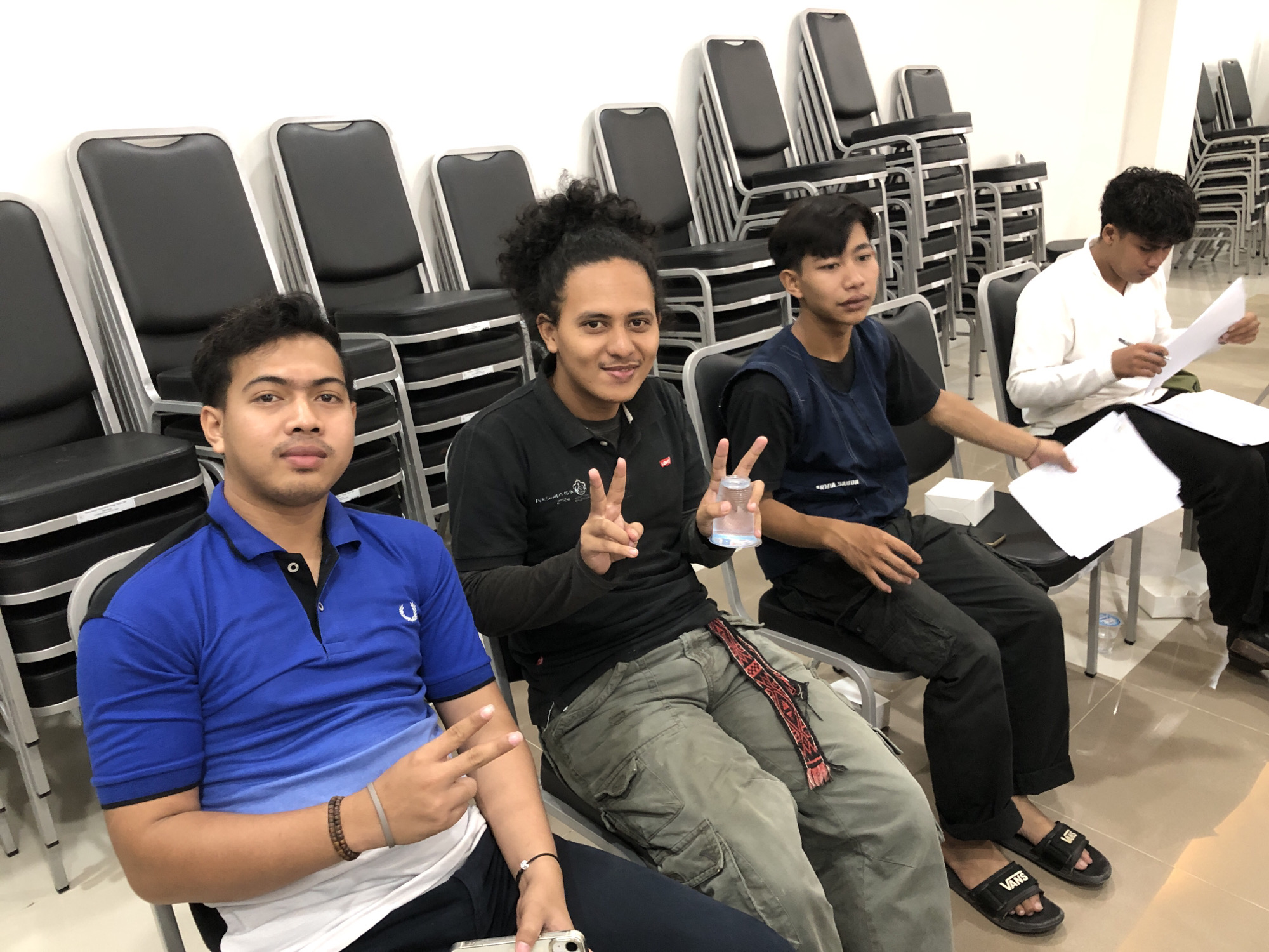 Four young adult males are seated in a room, with stacks of chairs in the background. The man on the far left wears a blue polo shirt and is looking forward, holding up two fingers. Next to him, a man with curly hair in a black polo shirt is smiling and also holding up two fingers, with a glass of water in his other hand. The third man from the left wears a blue vest over a dark shirt and is looking down at some papers. The man on the far right wears a white long-sleeved shirt and is also looking at papers, holding a pen. The room has tiled flooring and white walls. This scene appears to be captured within the vicinity of Lhokseumawe, Indonesia, given the context provided, potentially a classroom or meeting space.